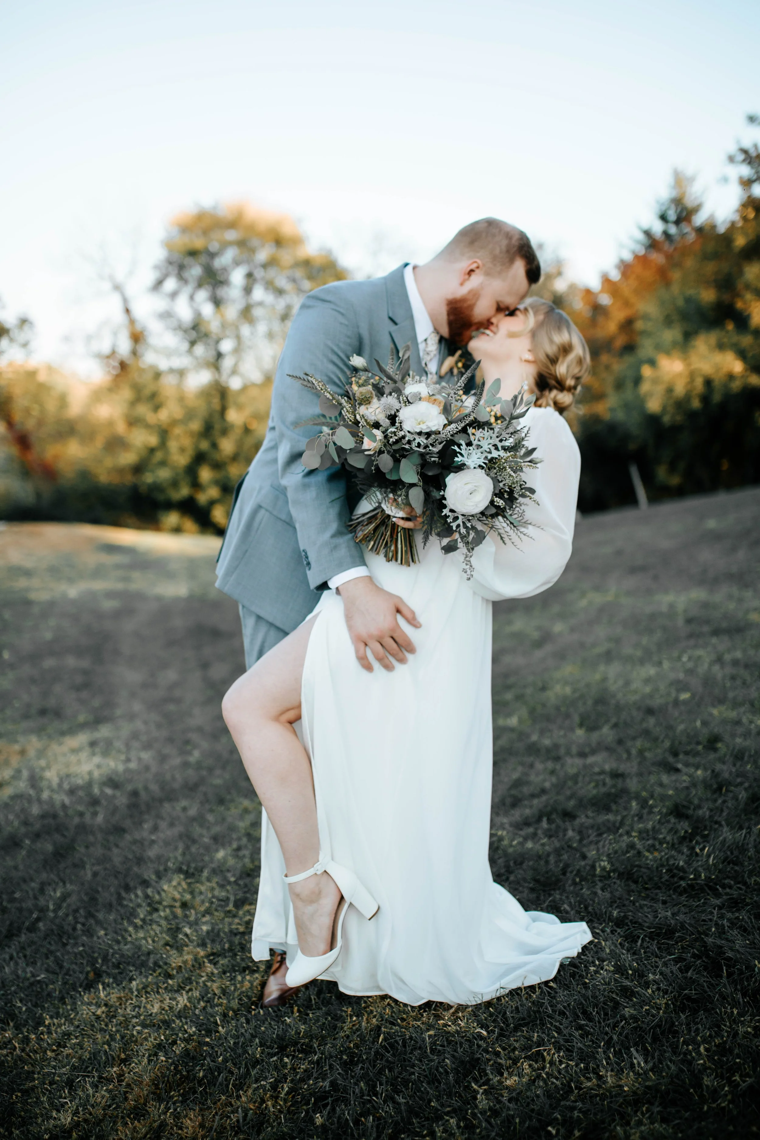 A newlywed couple shares a kiss outdoors during sunset, with the groom in a gray suit and the bride in a white dress holding a bouquet of flowers, surrounded by trees with autumn leaves.