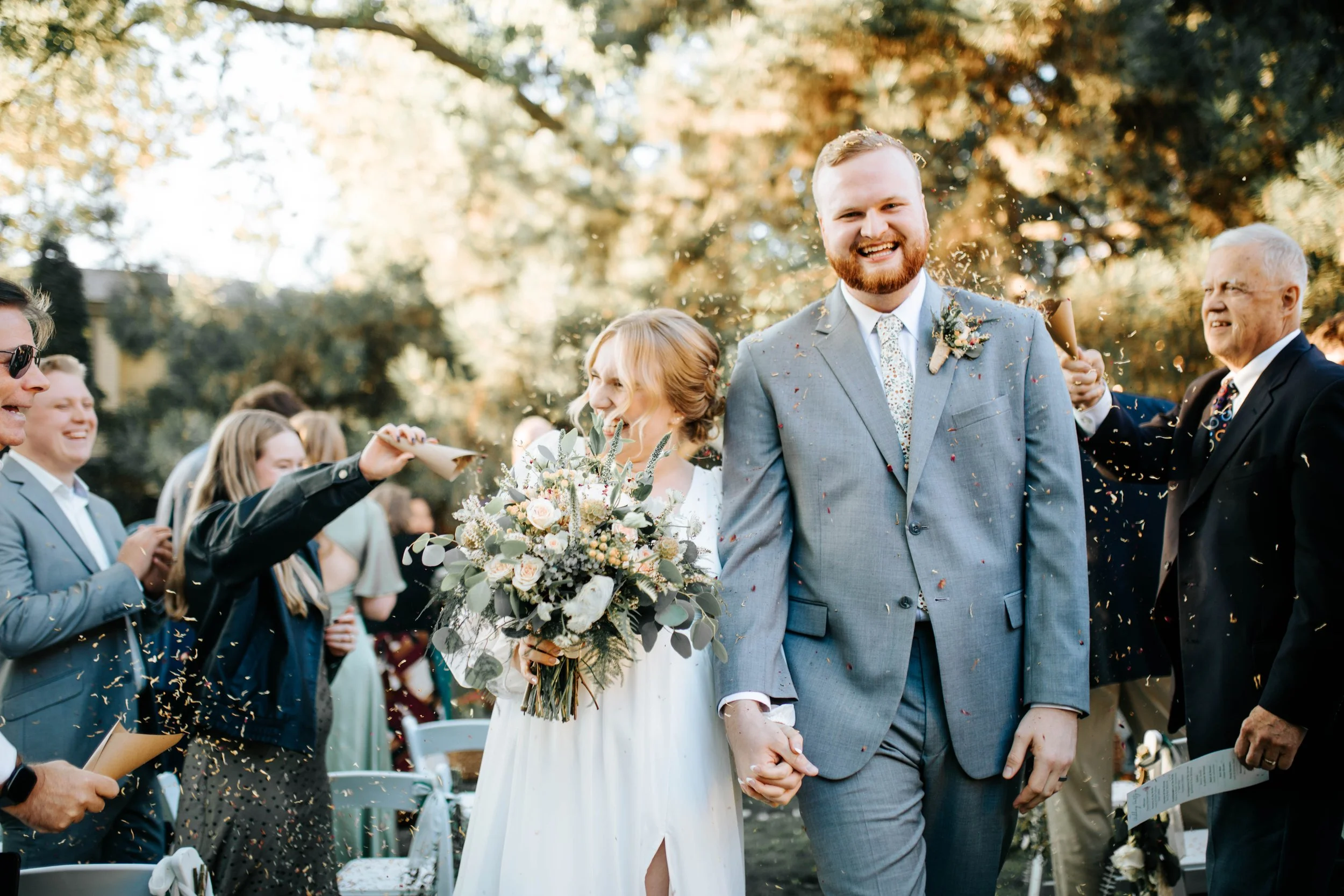 A newlywed couple walking hand-in-hand down an outdoor aisle, surrounded by wedding guests throwing confetti during daylight. The bride is holding a large bouquet of white and green flowers, and the groom is smiling in a grey suit, white shirt, and f