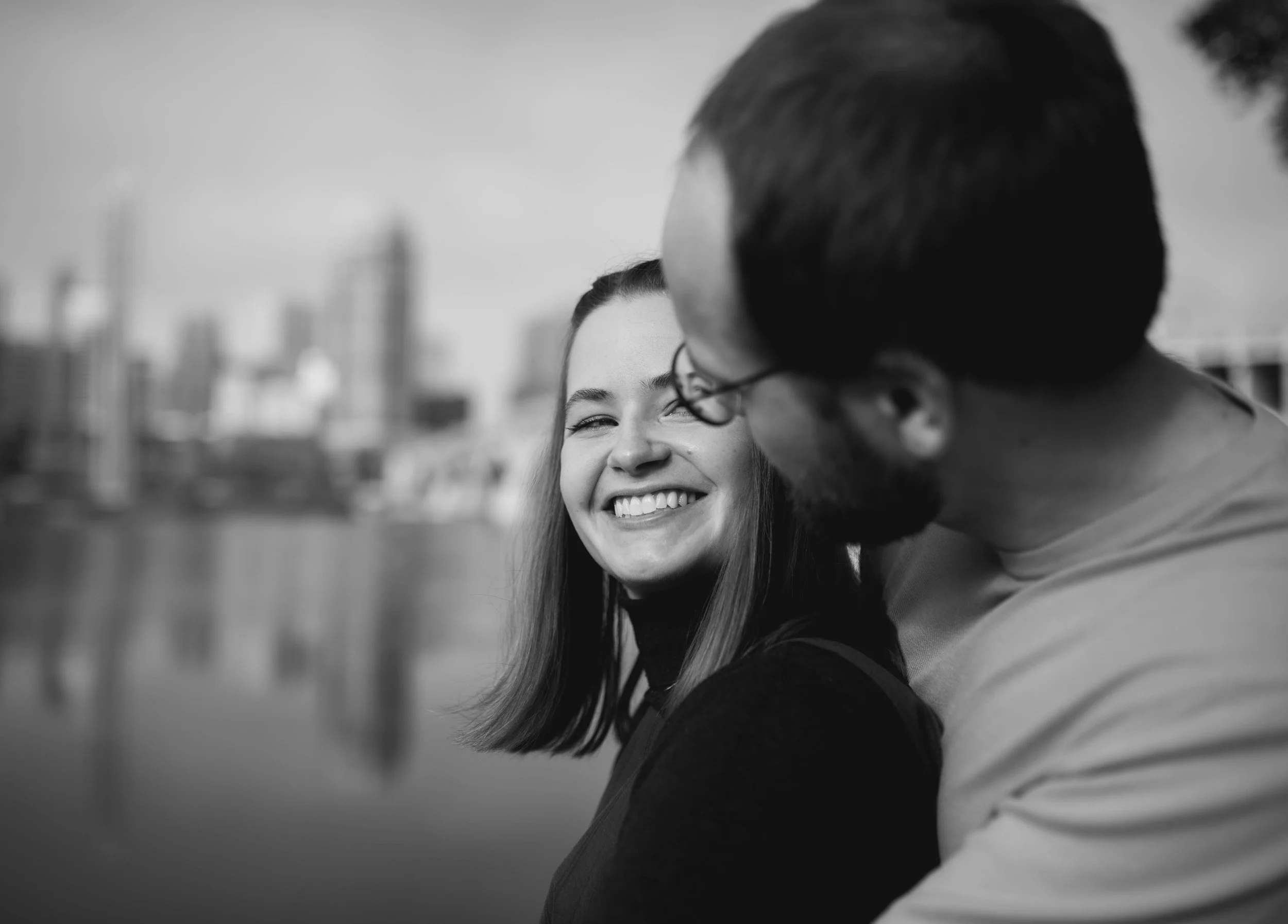 A black-and-white photo of a smiling woman and man, close together outdoors with city buildings in the background.