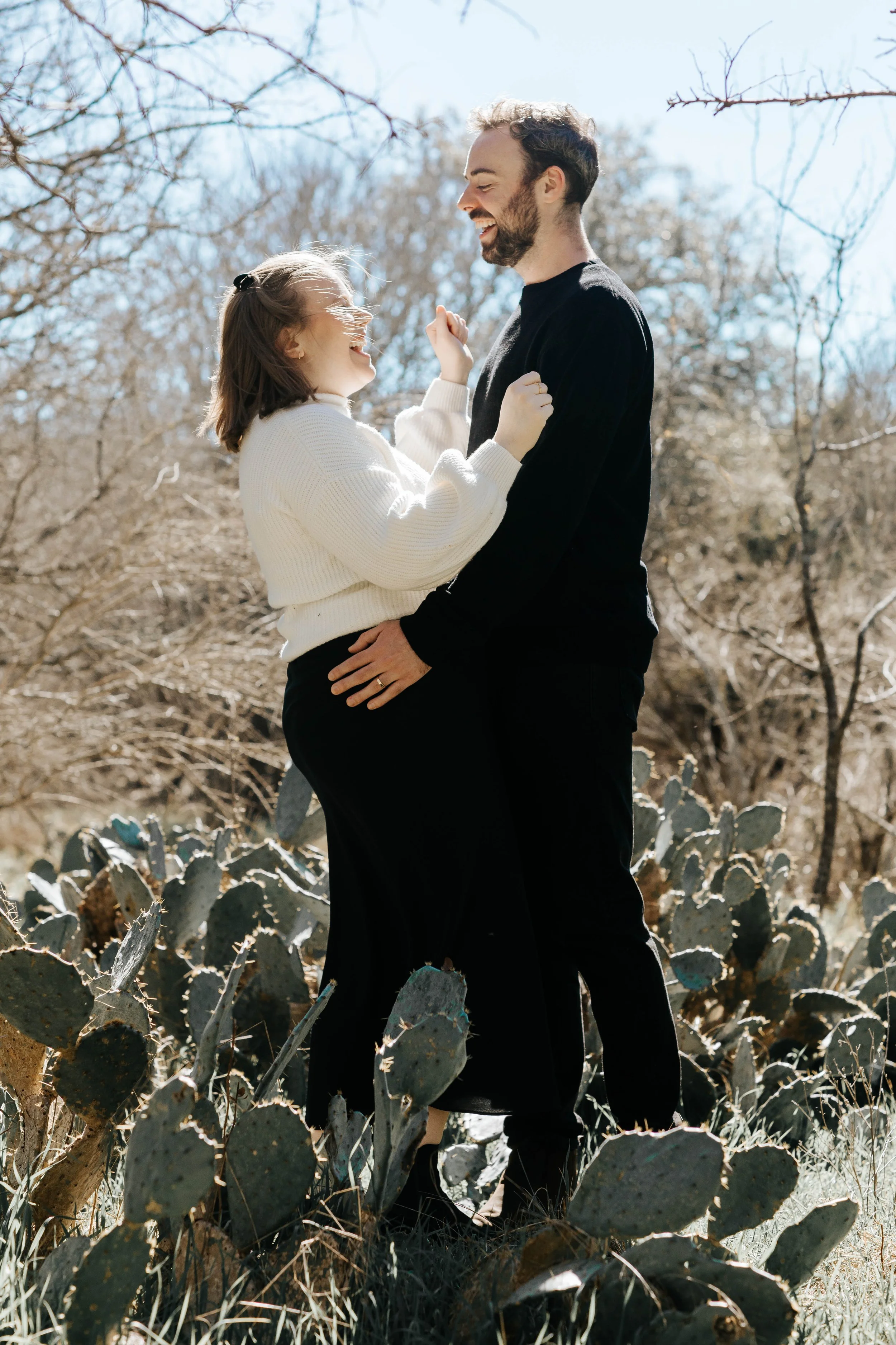A smiling man and woman are standing close together outdoors among cactus plants, enjoying each other's company on a sunny day.