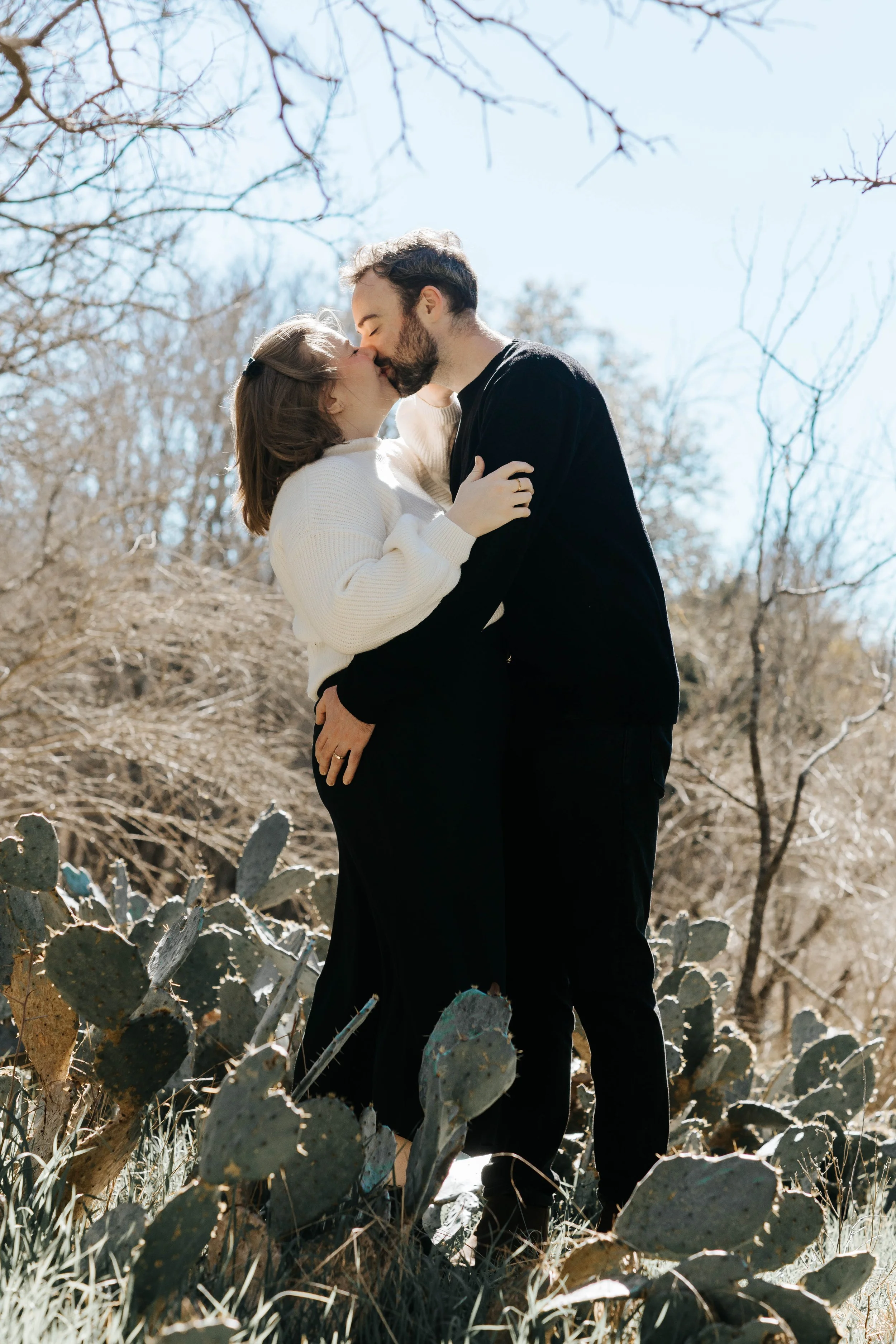 A couple sharing a kiss outdoors among cacti and leafless trees.
