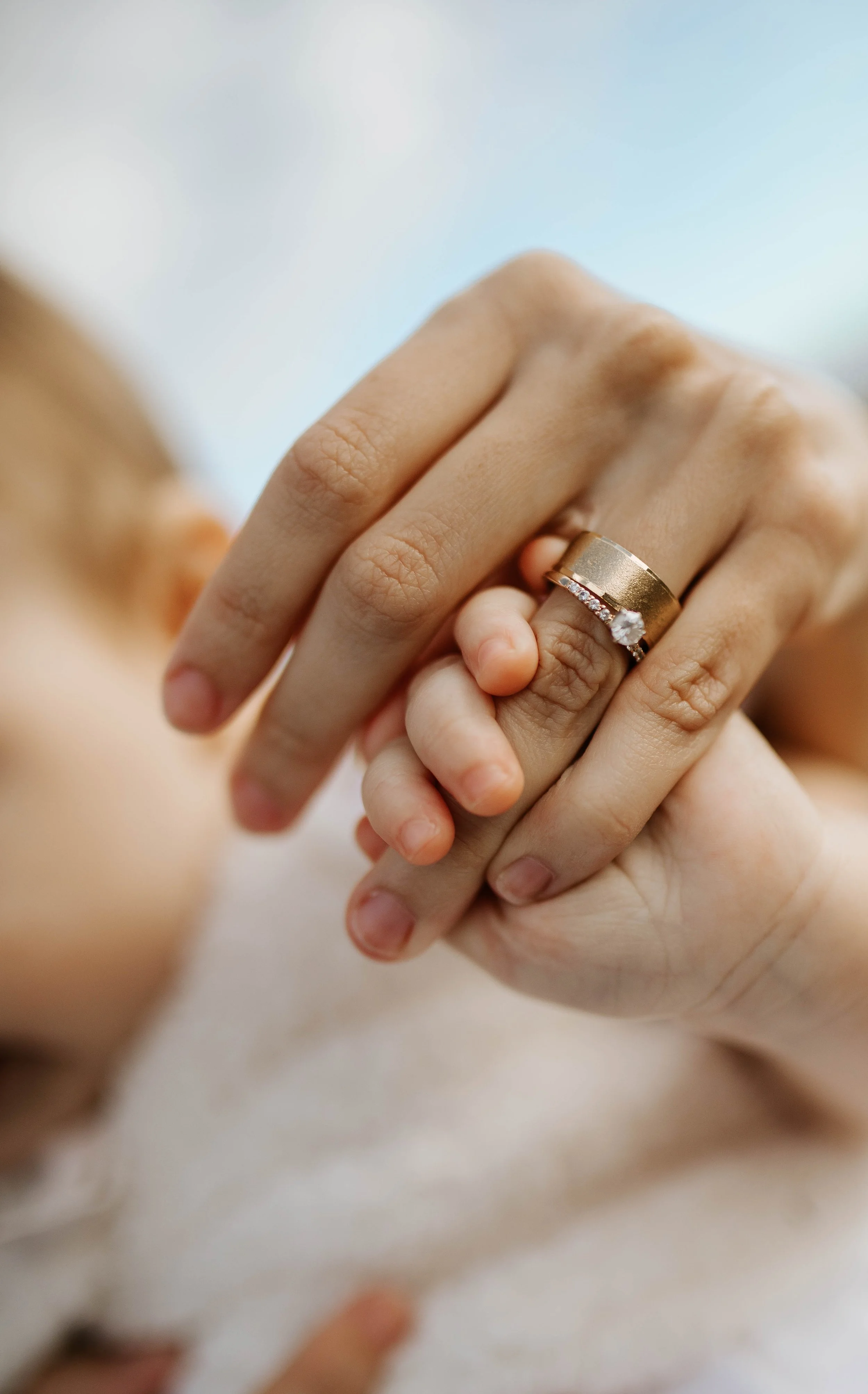 Close-up of an adult's hand wearing a gold wedding band with a diamond, holding a baby's hand.