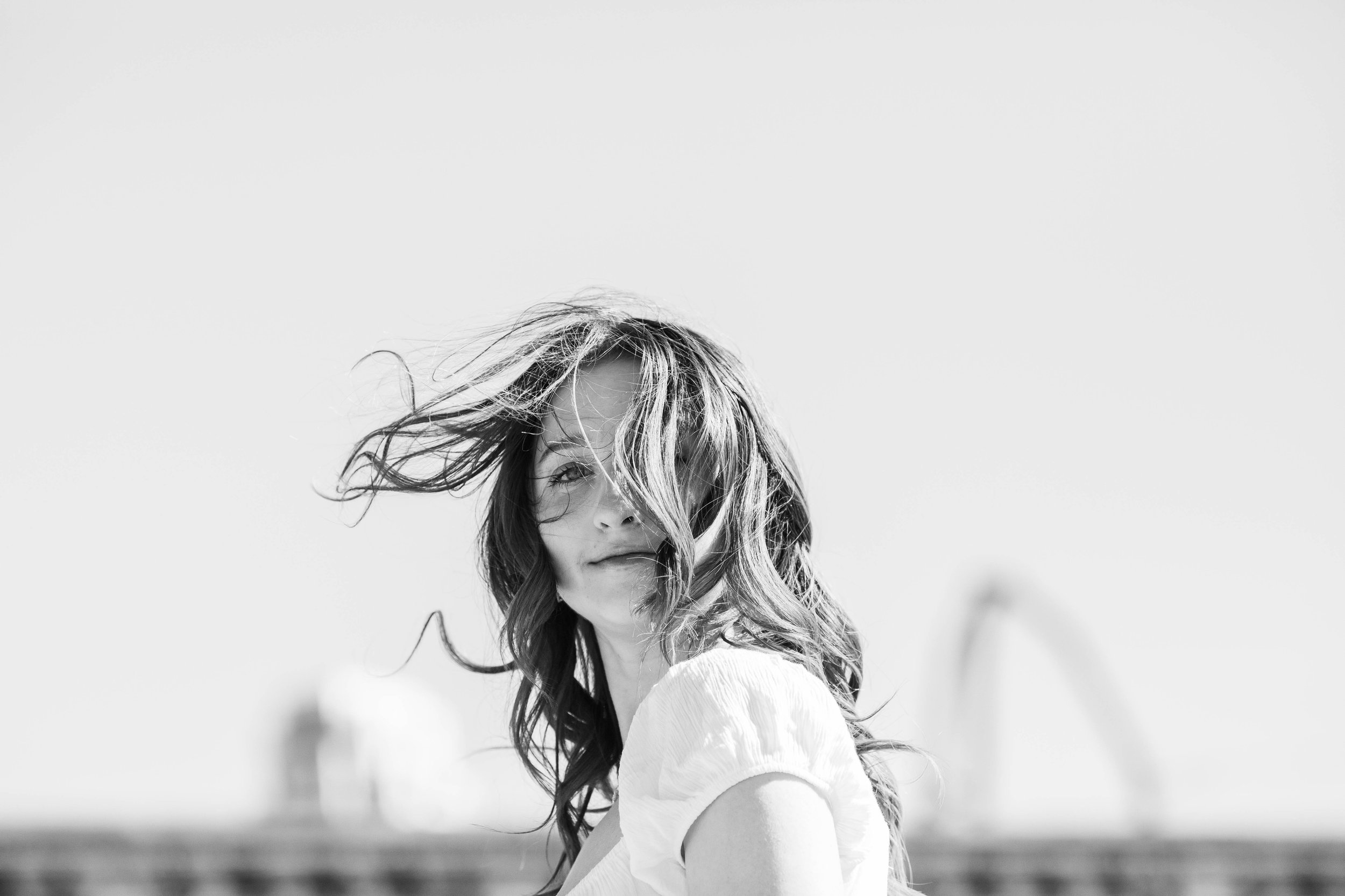 Black and white photo of a woman with wavy hair blowing in the wind, looking over her shoulder with a slight smile.