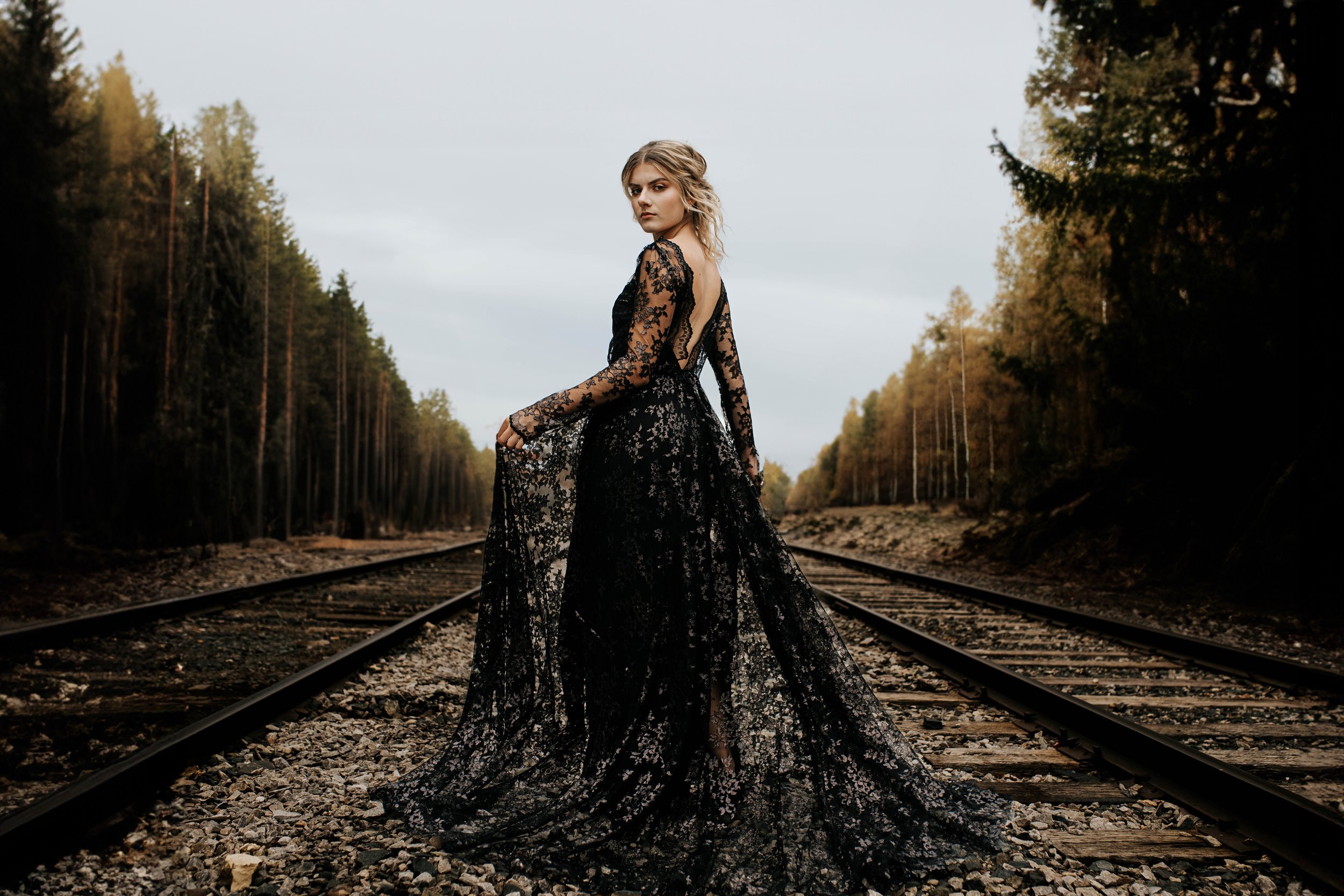 A woman in a long black lace dress standing on railroad tracks with a forest in the background.