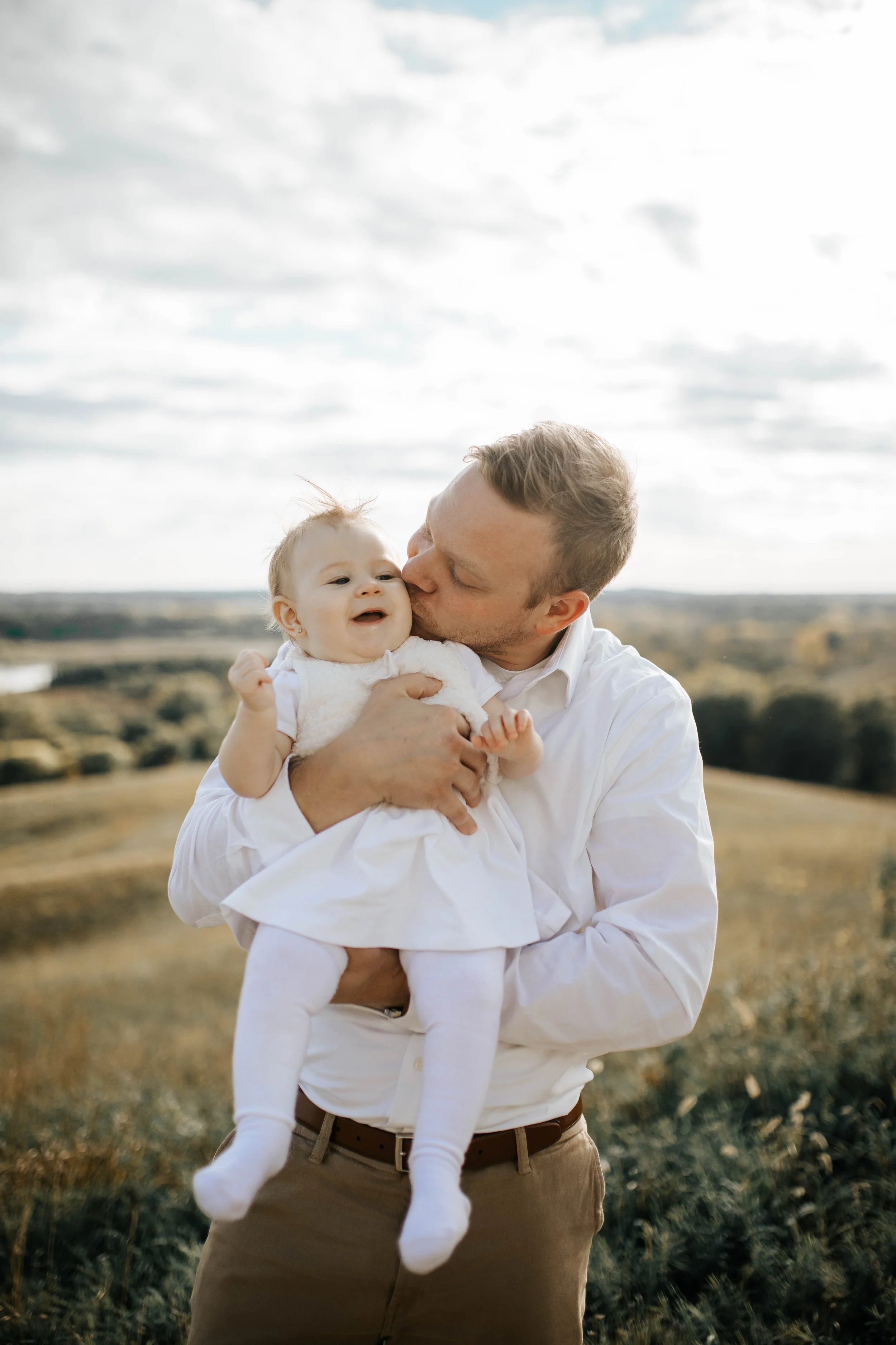 A man in a white shirt holding a crying baby girl dressed in white outdoors in a grassy field with cloudy sky.
