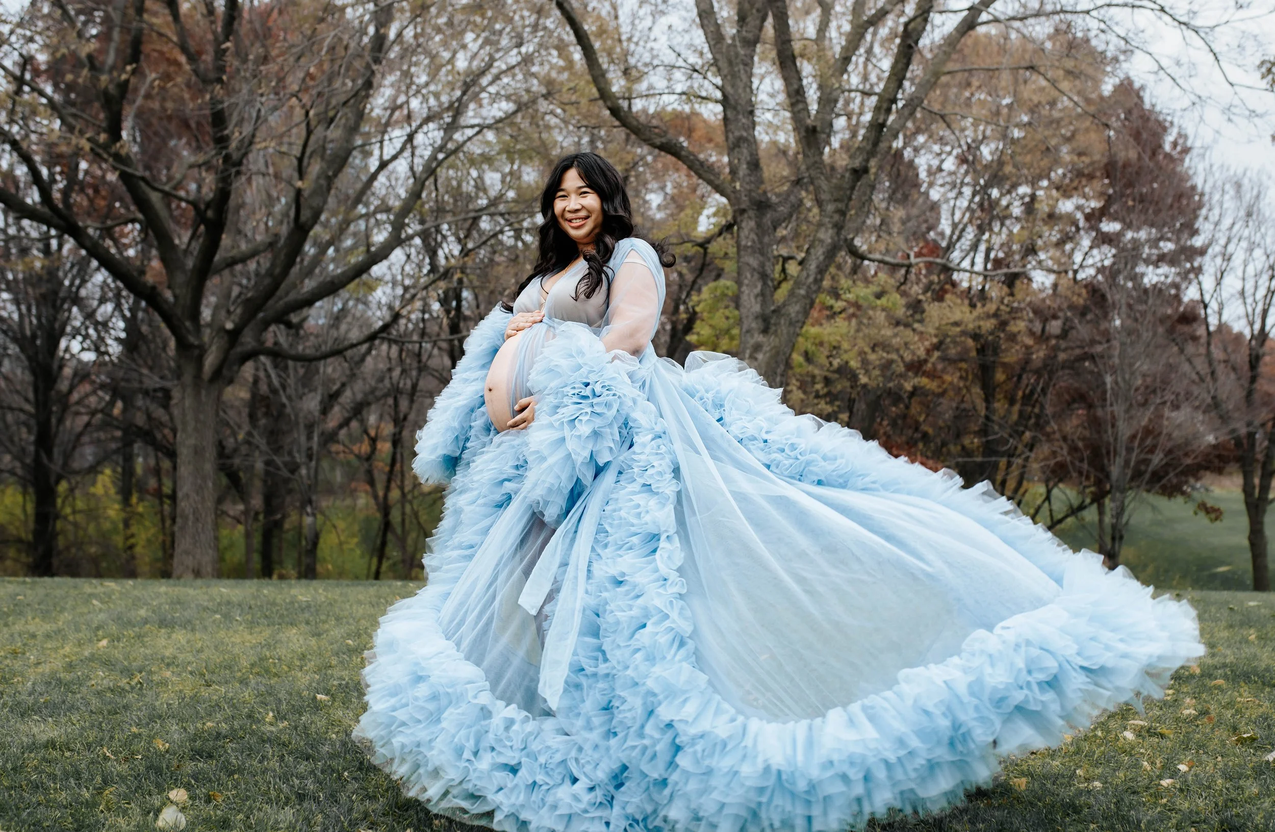 A pregnant woman wearing a flowing blue ruffled gown, smiling outdoors in a park with trees in the background.