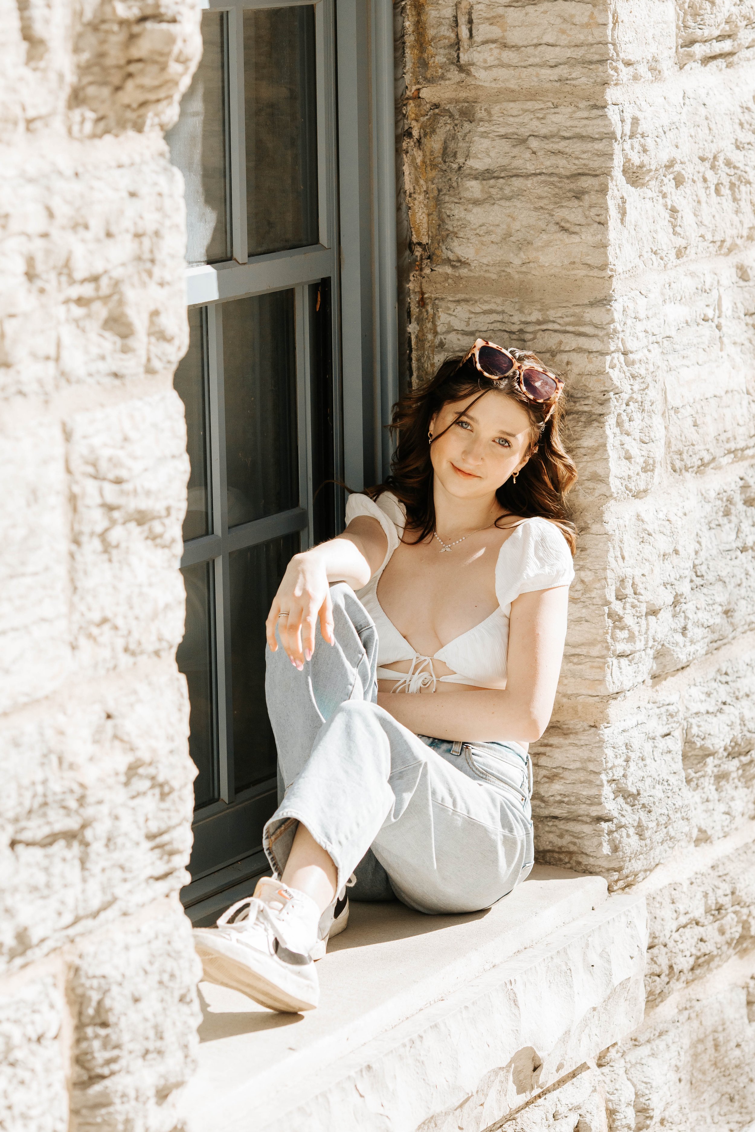 A woman sitting on a windowsill outside a brick building, wearing a white crop top, light gray jeans, white sneakers, and sunglasses on her head.
