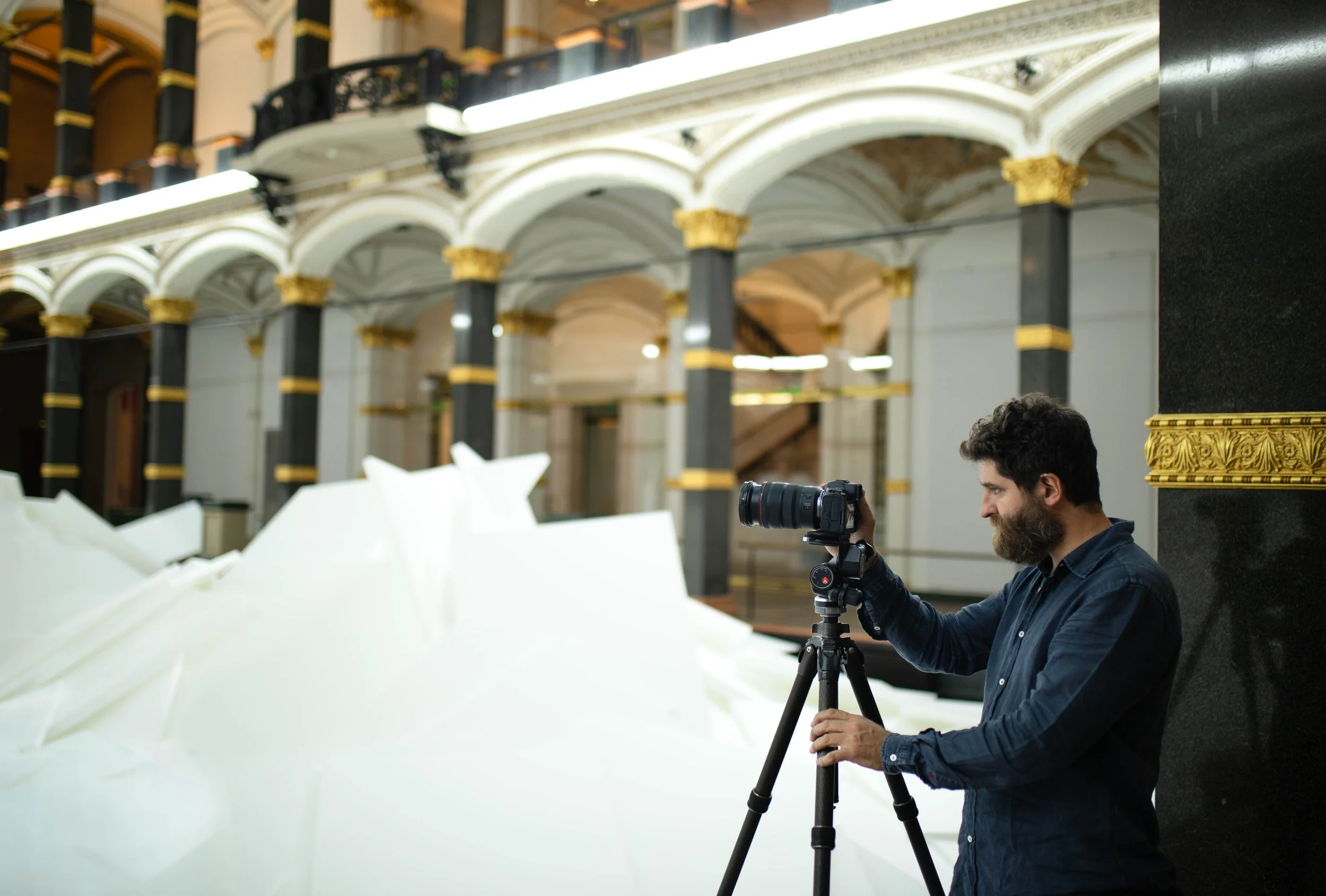 A man with dark hair and a beard operating a professional camera on a tripod inside a grand building with ornate architecture and white draperies.
