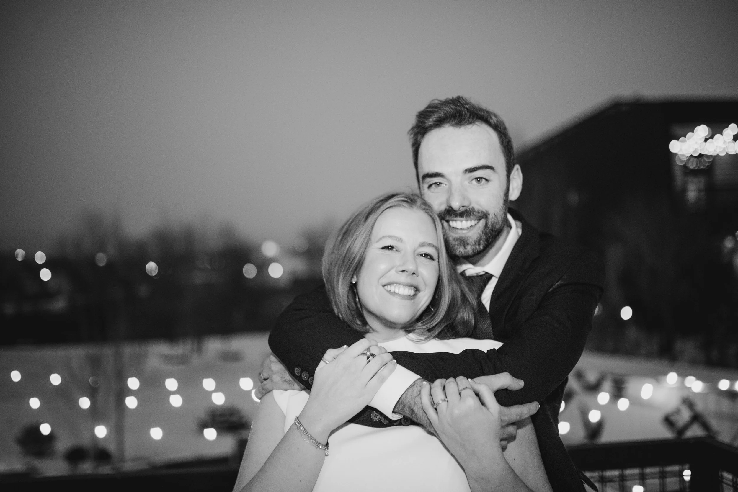 A black-and-white photo of a happy couple embracing outdoors at night, blurred city lights in the background.