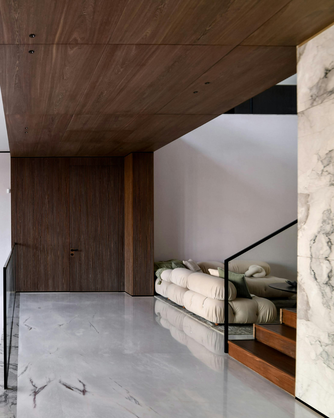 Modern interior with marble flooring, wooden ceiling, and a beige couch near stairs with black railing.