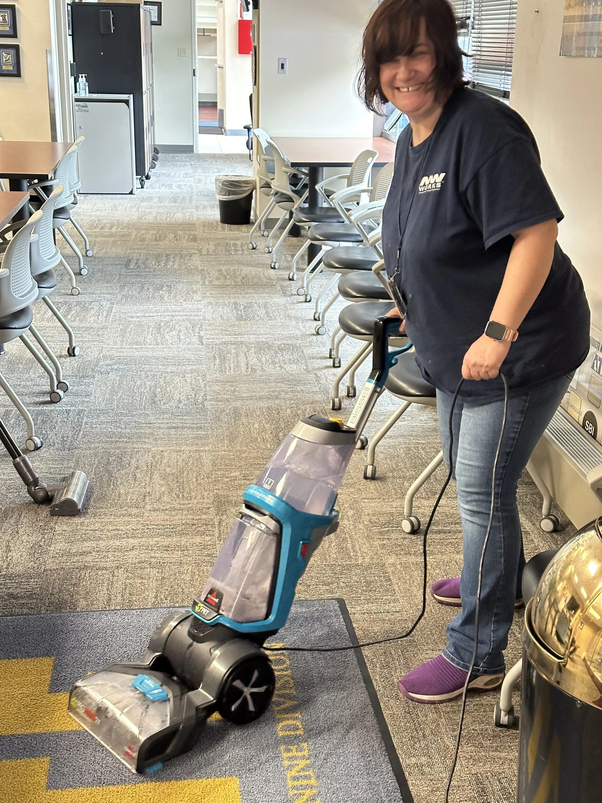  A middle aged woman is shown wearing a navy blue NW Works t-shirt and smiling at the camera as she vacuums. She is vacuuming in a carpeted office space with tables and chairs. 