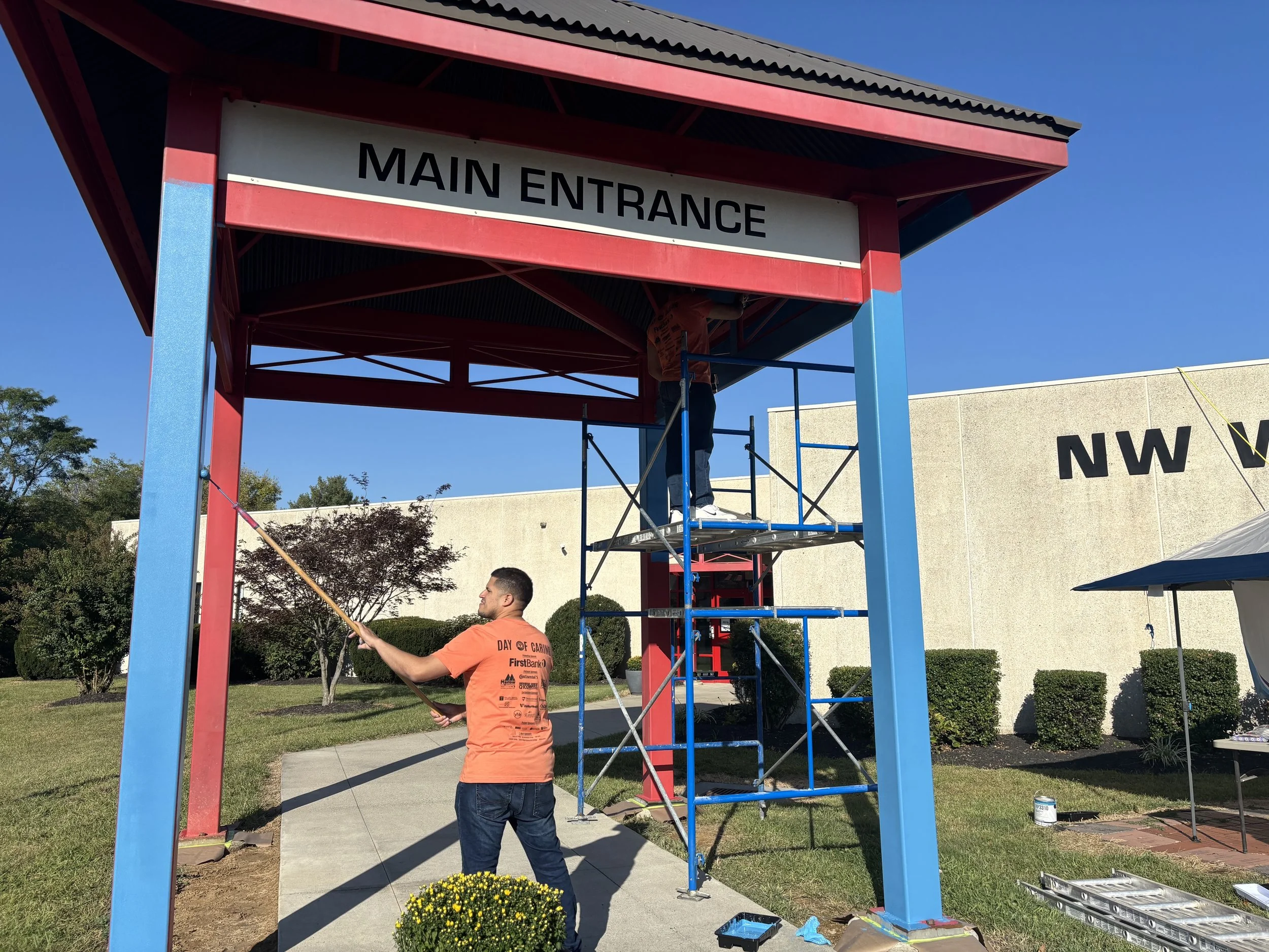A man in an orange t-shirt is shown painting with a roller brush. The man is painting the main entrance of NW Works in blue to cover up the red formerly used around the building.