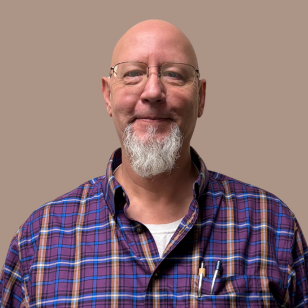 An older caucasian man with a gray beard and glasses named Troy Hollon, is smiling. Troy is wearing a purple, tan, blue and white collared shirt.