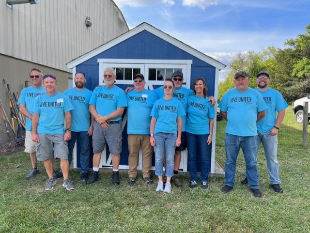  10 volunteers from Continental stand in front of their completed blue shed in the NW Works Garden. They are all wearing blue "Live United" shirts for the 2023 United Way Day of Caring. 