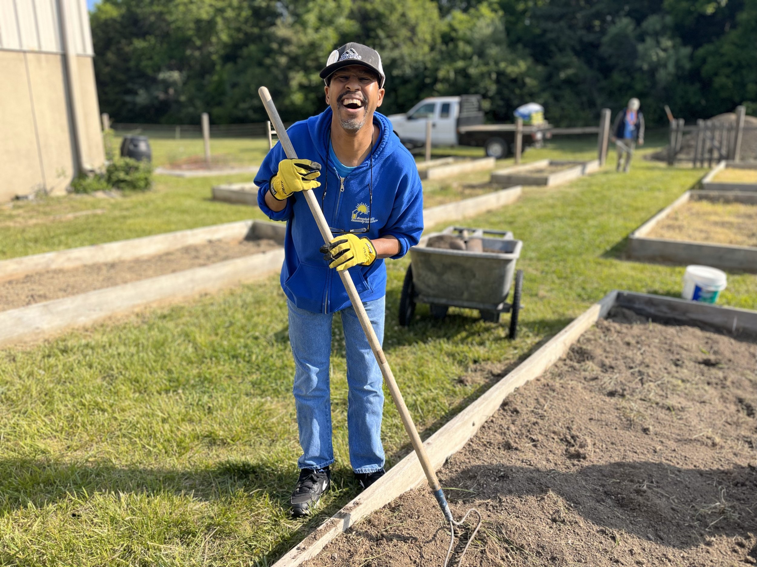  A man in a blue sweatshirt and a hat stands with a rake in his hand. A huge smile is across his face and he is leaning toward the camera. He is in the NW Works garden clearing a raised garden bed and spreading soil. 