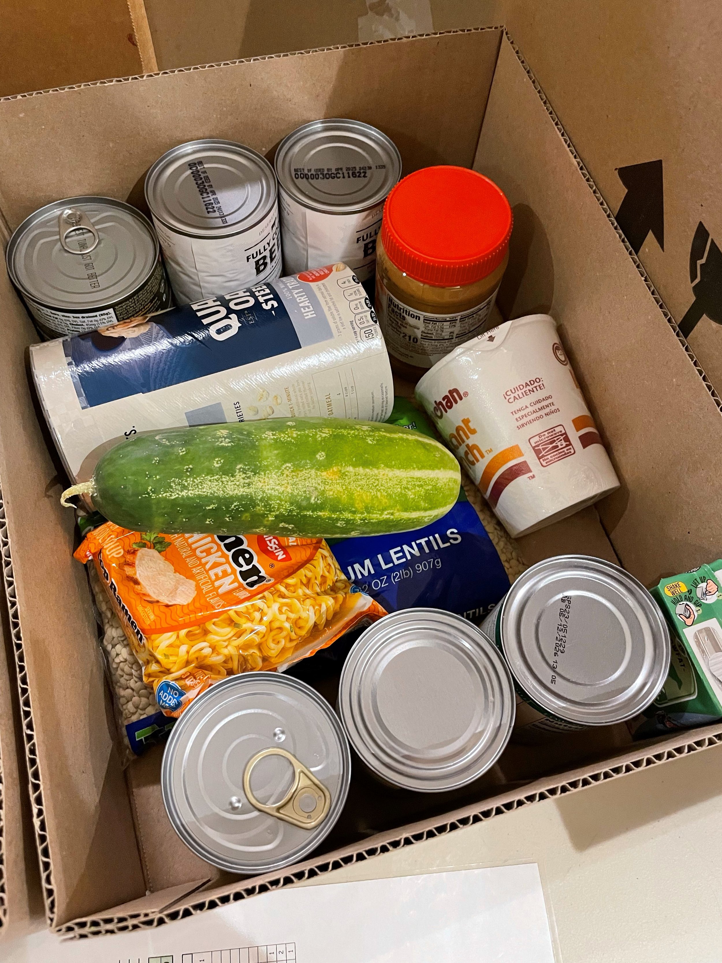  A cardboard box full of food donations is shown. The donations include several cans, peanut butter, ramen, oatmeal, lentils and a cucumber from the NW Works Garden. 