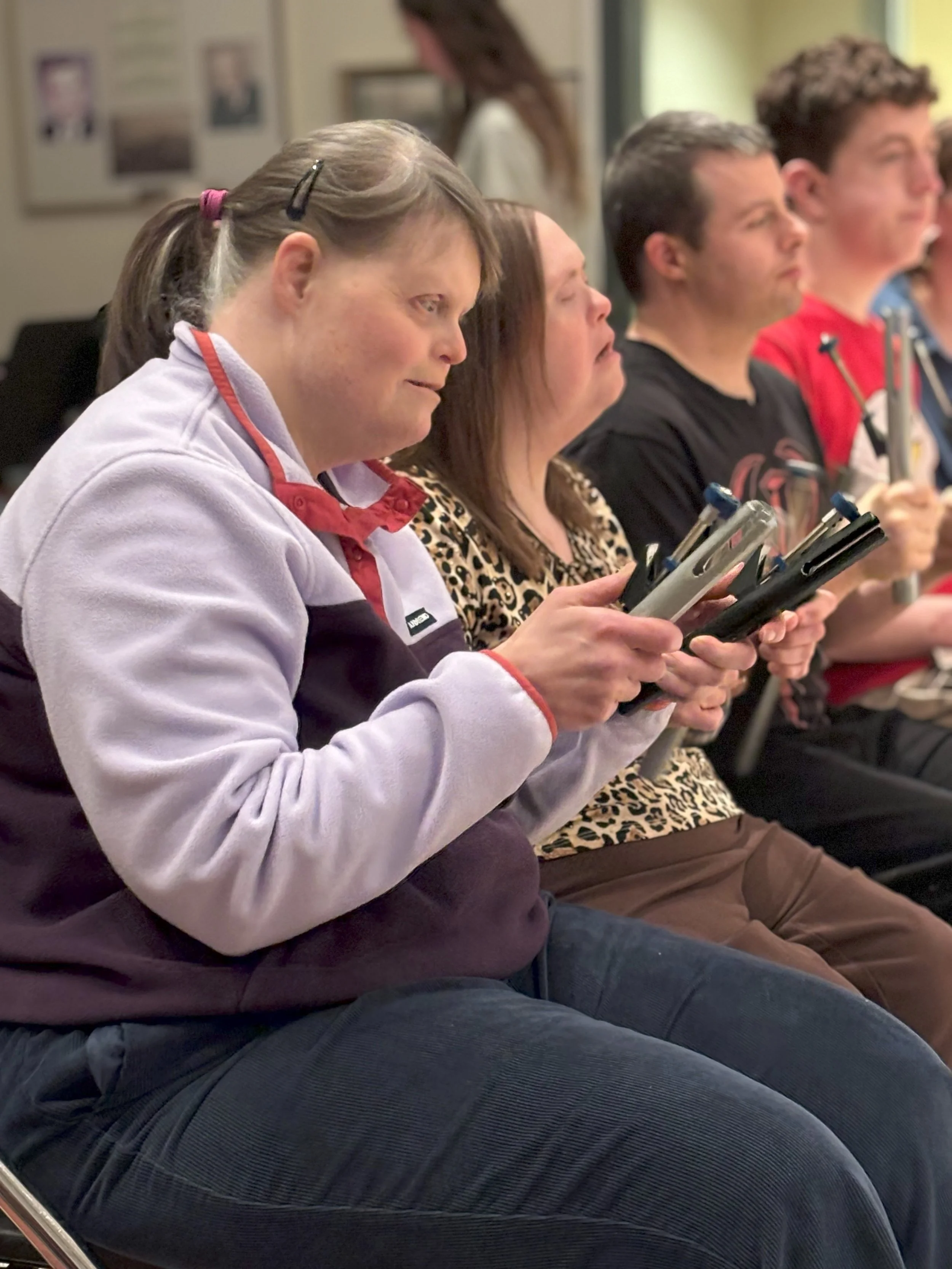  Two women sit in chairs during the Chimers rehearsal. They are focused on the conductor and are holding their chimes. 