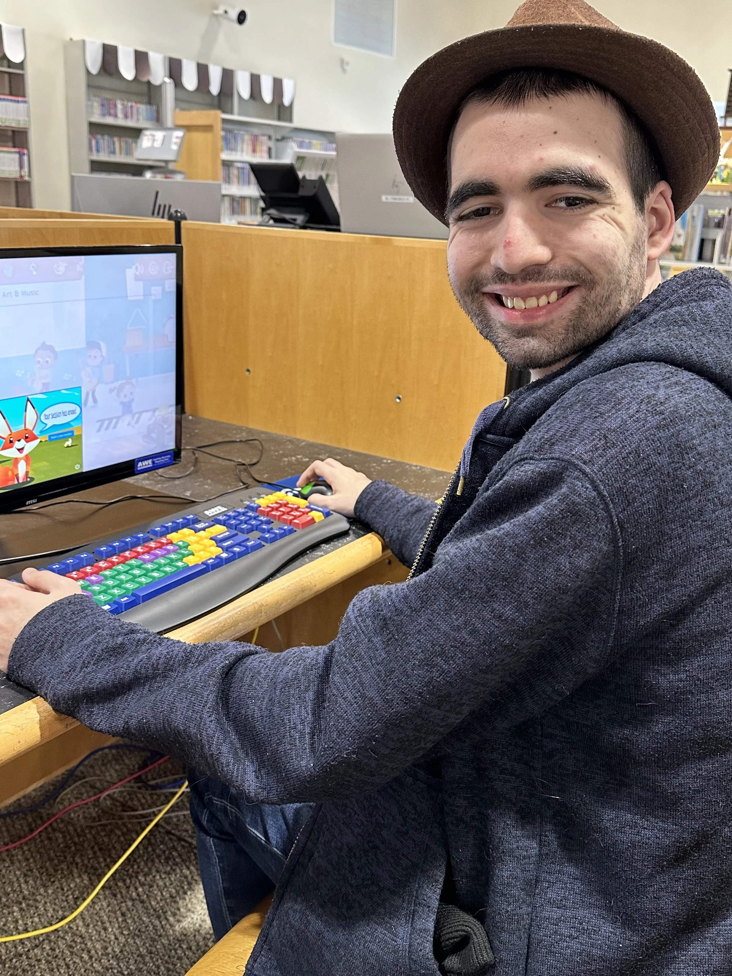  A man sits at a computer desk and is turned to have his picture taken. He is wearing a brown fedora and smiling. 