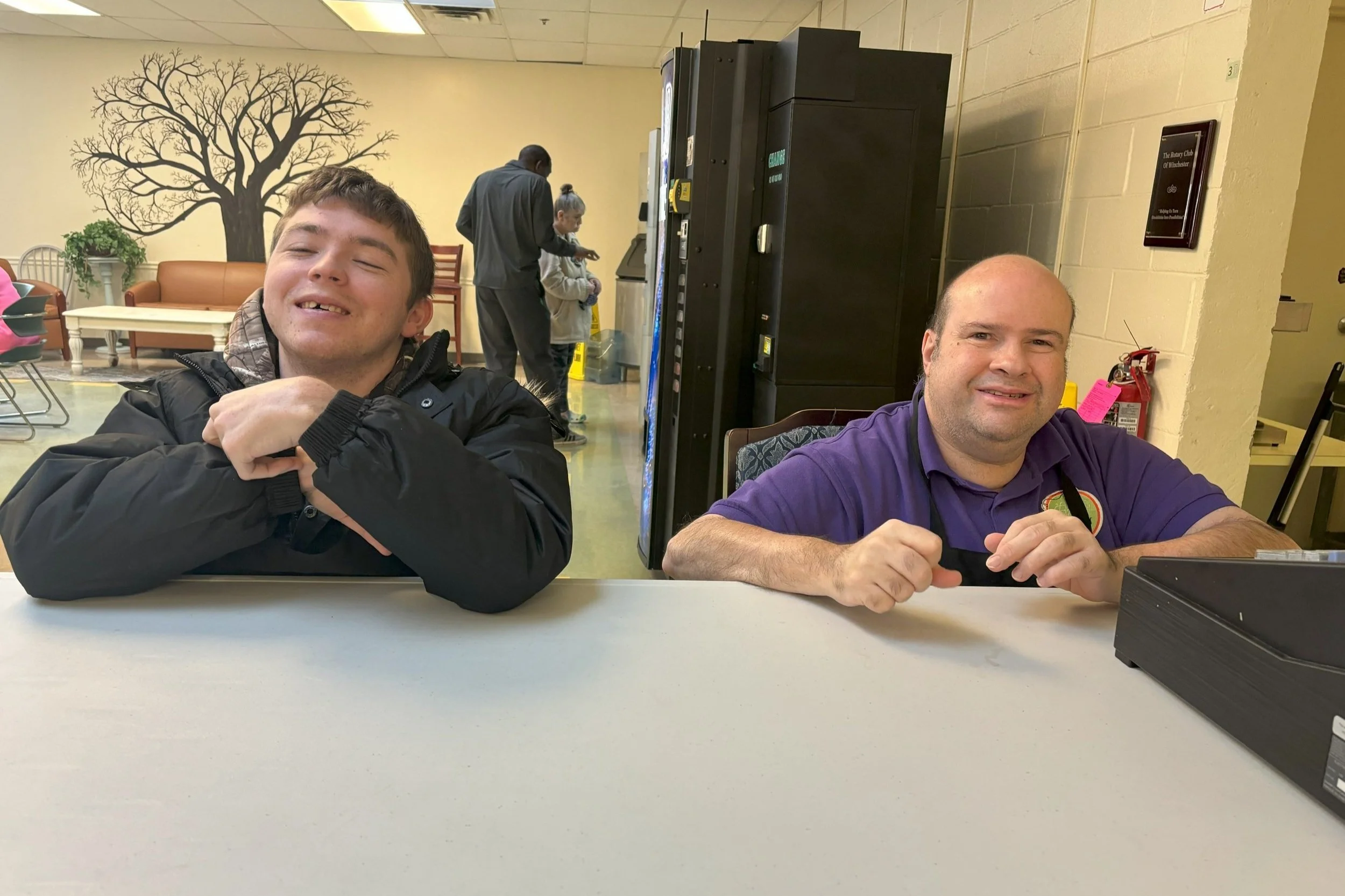  Two men sit behind a table in the Career Cafe at NW Works. They are beside a cash register and are serving as cashiers for the day. 