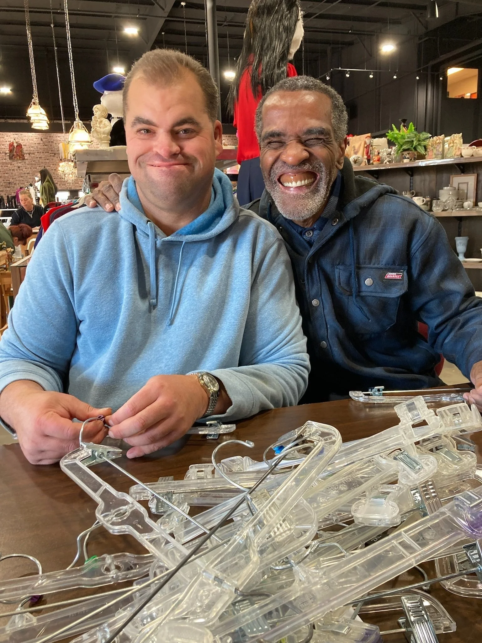  Two men sit at a table and they are touching shoulders. Both men are wearing blue and they are smiling at the camera. 