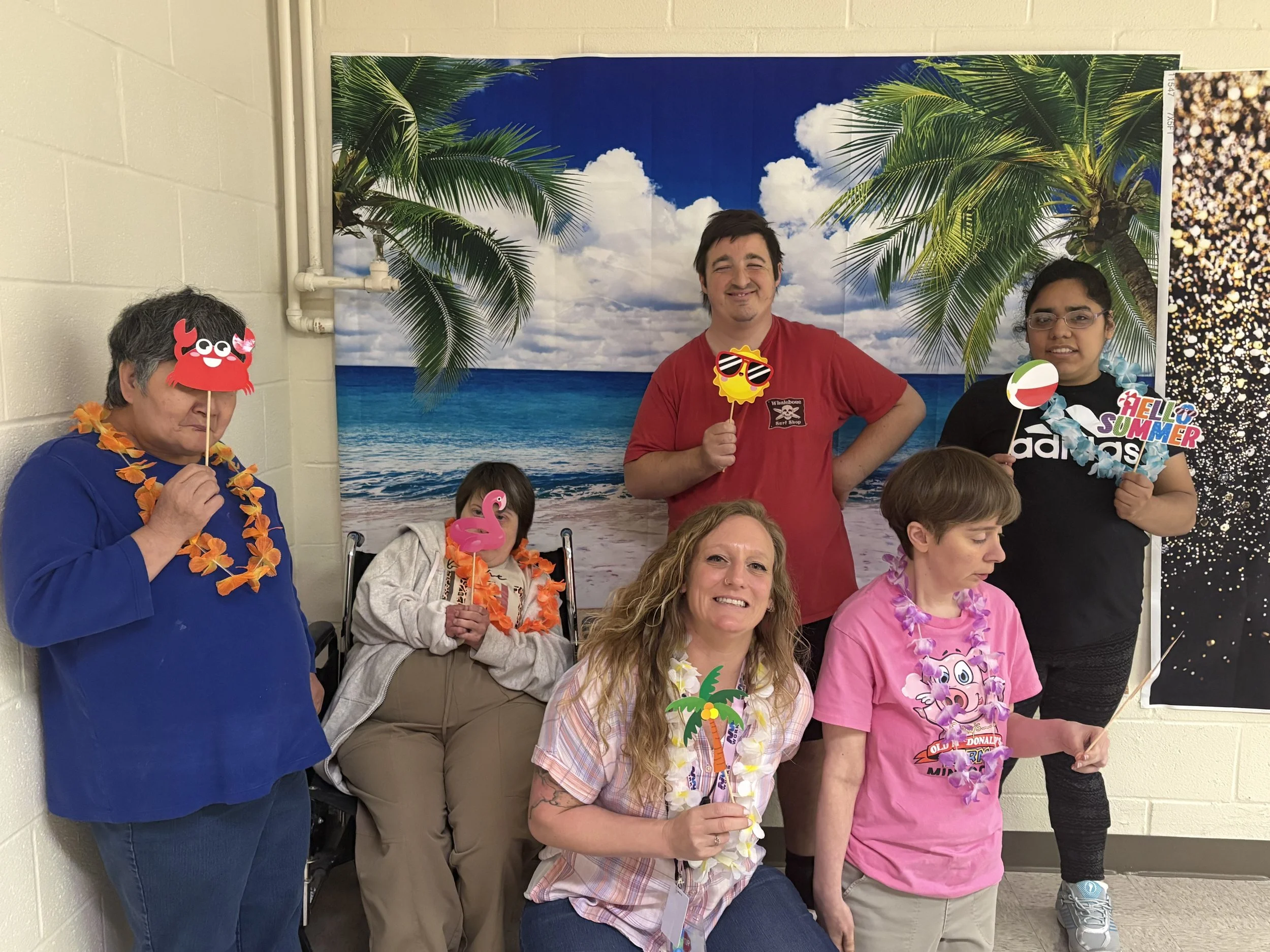  A group of participants at NW Works are shown smiling in front of a beach scene at a selfie station. The people are holding up various summer themed props. 