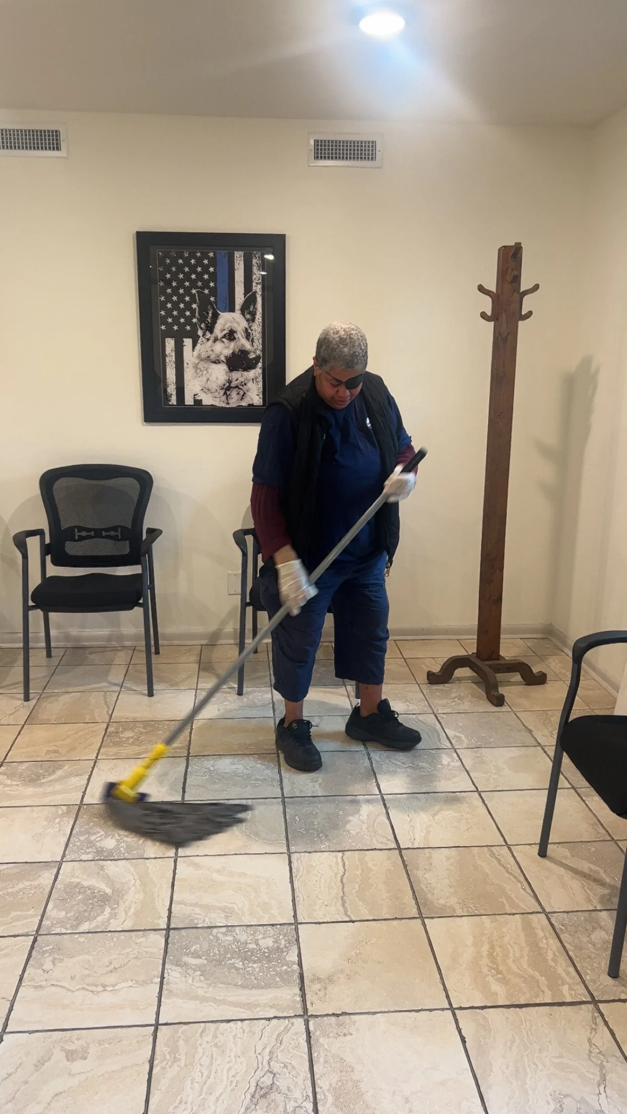  A middle aged woman stands in the middle of a white tile floor with a mop. She is in the process of cleaning the floor. Behind her is a coat rack, some chairs, and a photo of a German Shepherd with a U.S. flag background. 