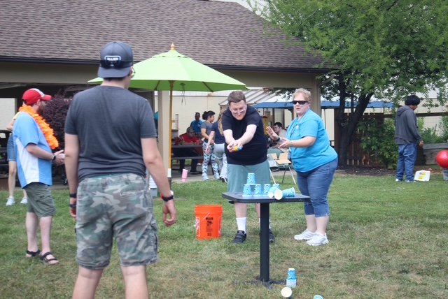  Several people stand around a table that has cups stacked in a pyramid foundation on it. There is a middle aged man trying to knock down the cups from a distance, while a staff member helps him and others watch. 