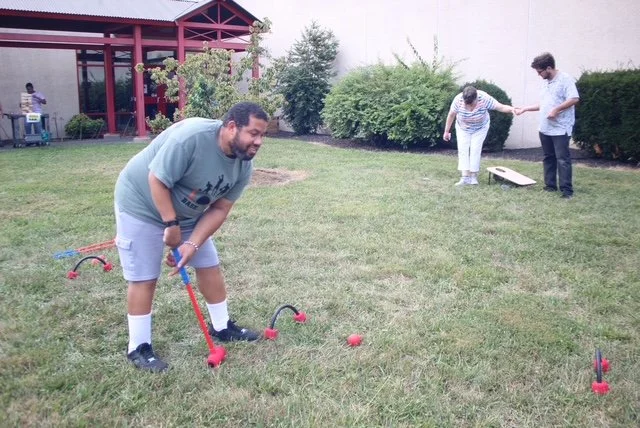 A man stands and is slightly bent over to play a game of croquet. He is holding the mallet with both hands. In the distance are two individuals preparing to play cornhole. 