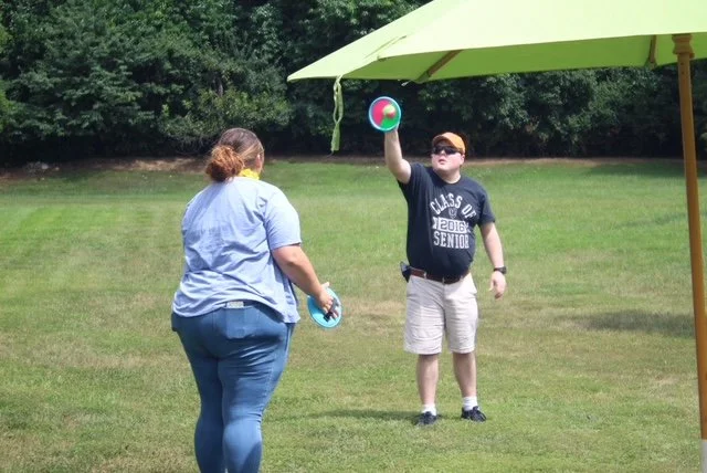  A woman has her back turned to the camera and a man is shown with his body toward the camera. They are throwing a ball back and forth and catching it with round velcro boards. 