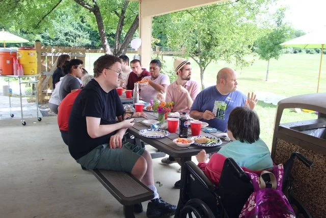  A large group of people sits at a table and watches others play yard games which is out of view of the camera. Everyone has a plate and drink in front of them. 