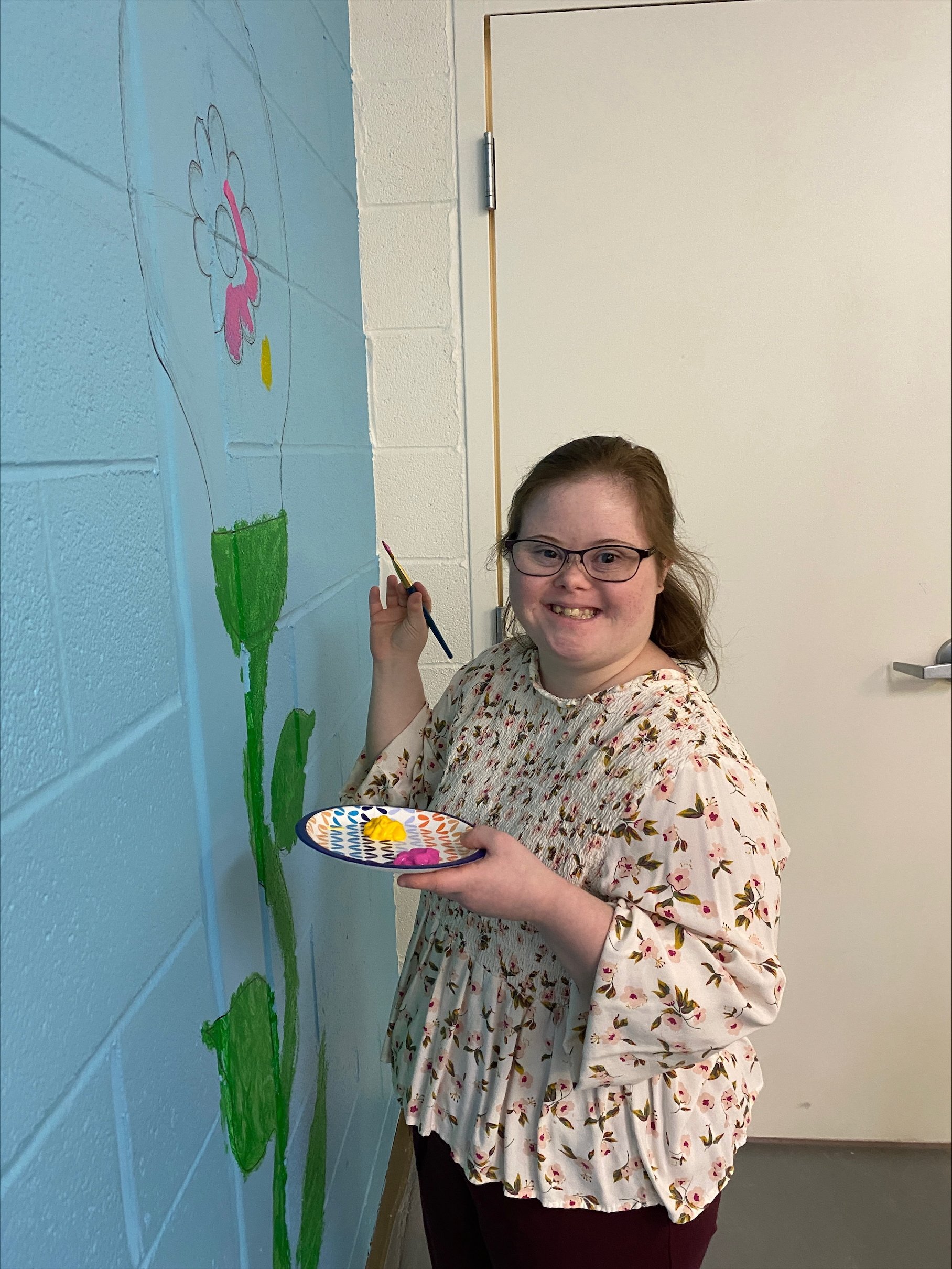  A young woman is shown smiling at the camera as she paints with pink and yellow paint on a flower with a lightbulb for a bloom in the Digital Literacy lab at NW Works. 