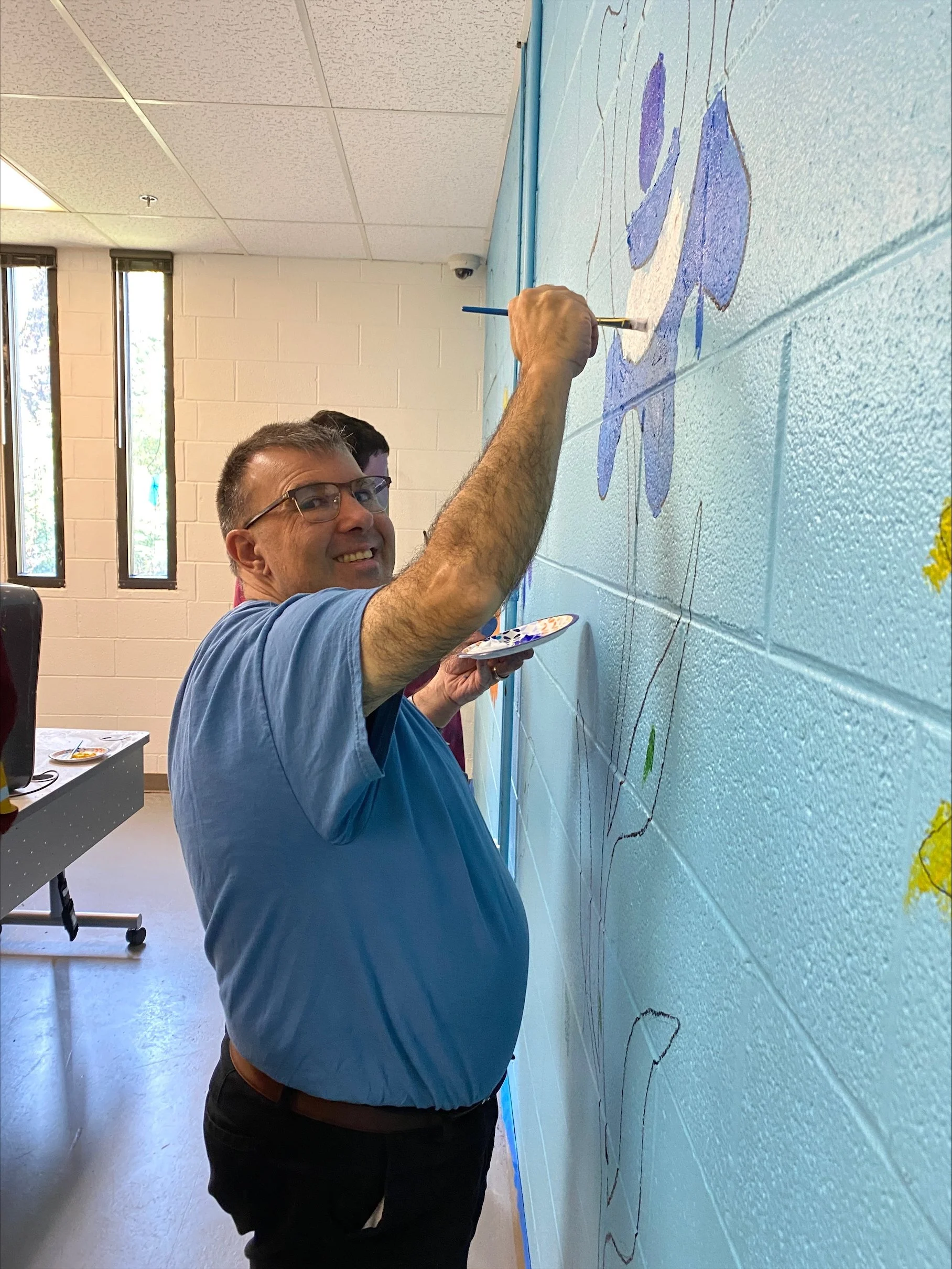  A middle aged man is shown smiling at the camera as he pauses mid-paint. The man is painting a blue and white flower on the wall in the Digital Literacy lab at NW Works. 
