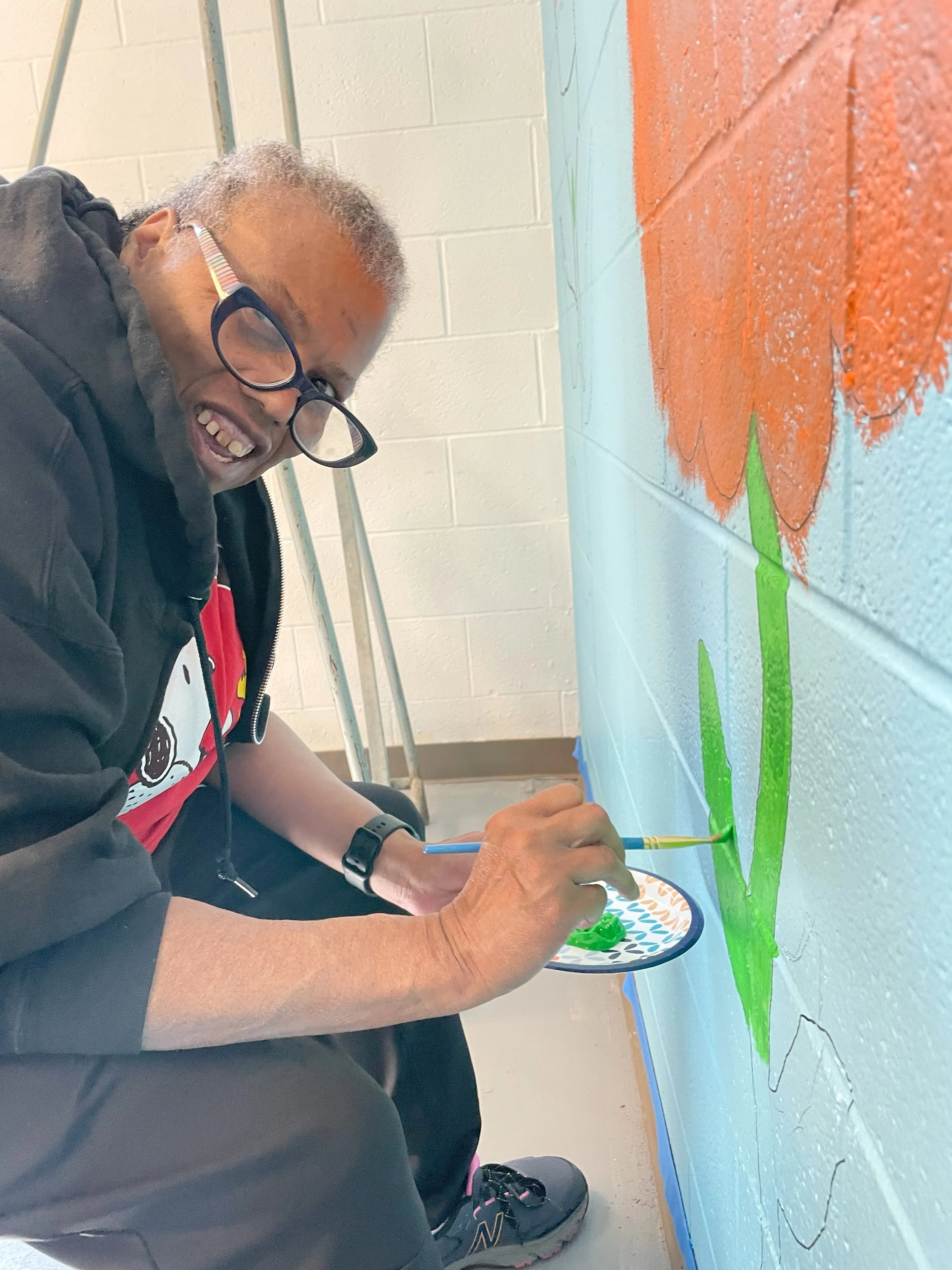  A middle aged woman is shown smiling and slightly hunched over as she paints the lower part of a stem on a flower on the wall of NW Works’ Digital Literacy lab. 