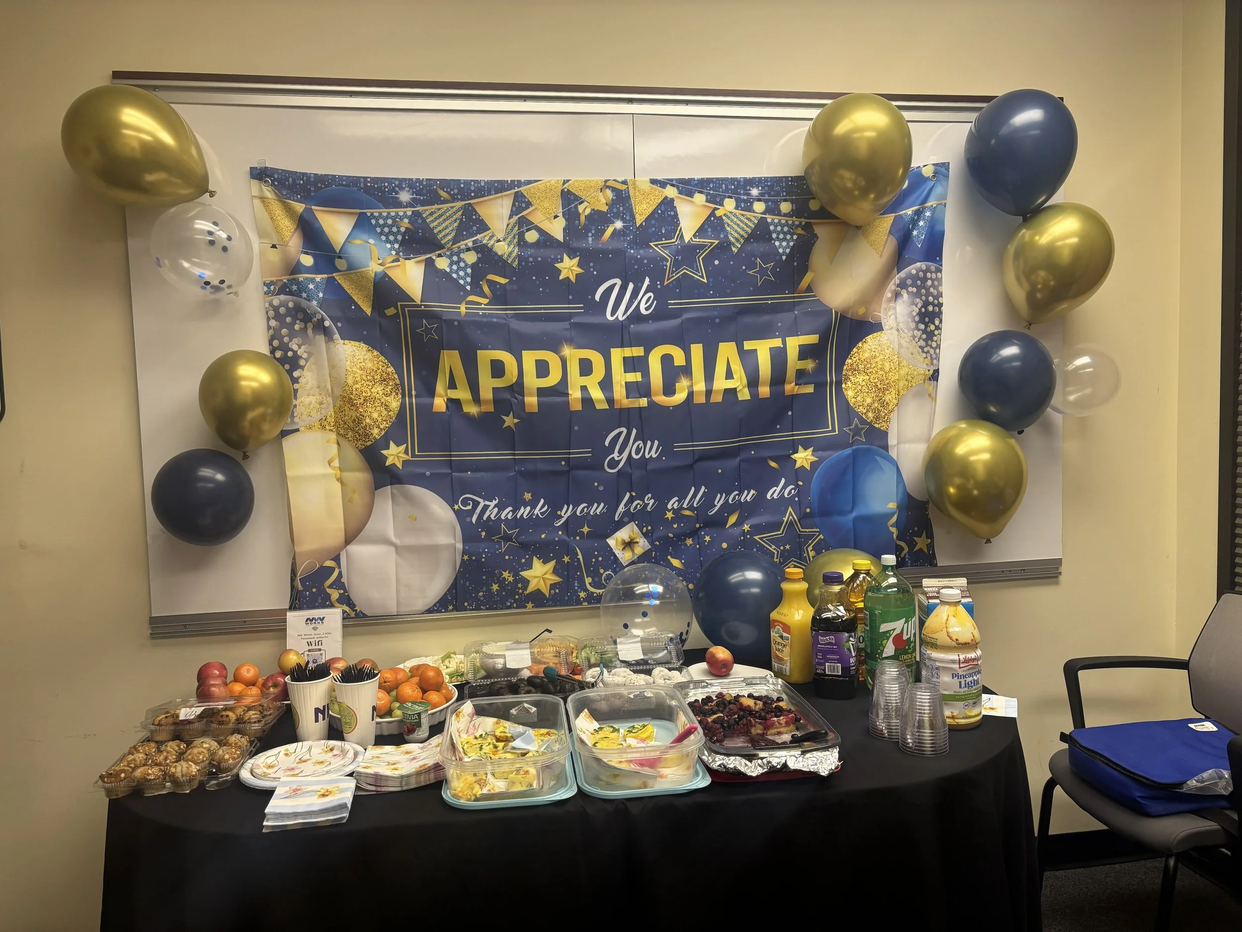  An Employee Appreciation banner with balloons is shown over a spread of breakfast foods and drinks. The banner and balloons are blue and gold, and the banner reads "We appreciate you. Thank you for all you do!" 