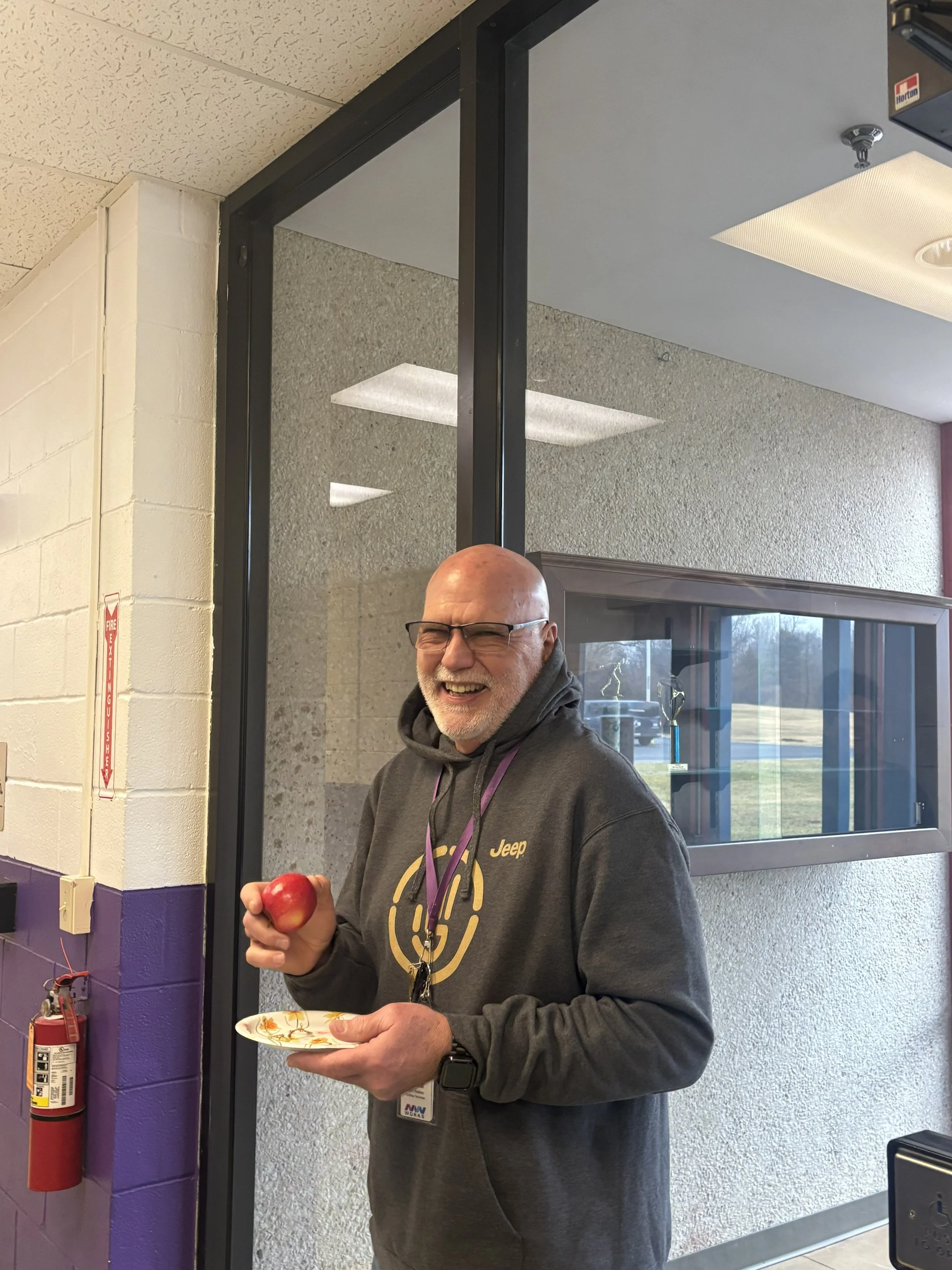  A middle aged man is shown smiling as he holds up a red apple in one hand and his plate in the other. The man is standing against a large glass window. 