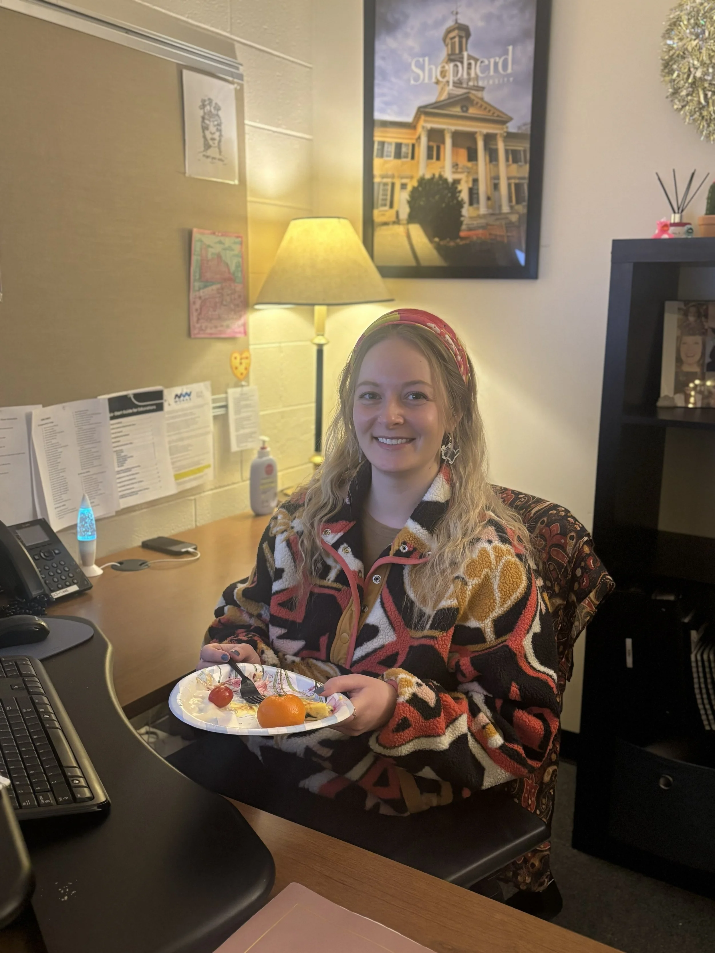  A young woman is shown holding up her plate of food as she sits at her desk. The woman is wearing a colorful patterned jacket in a warmly lit office space. 