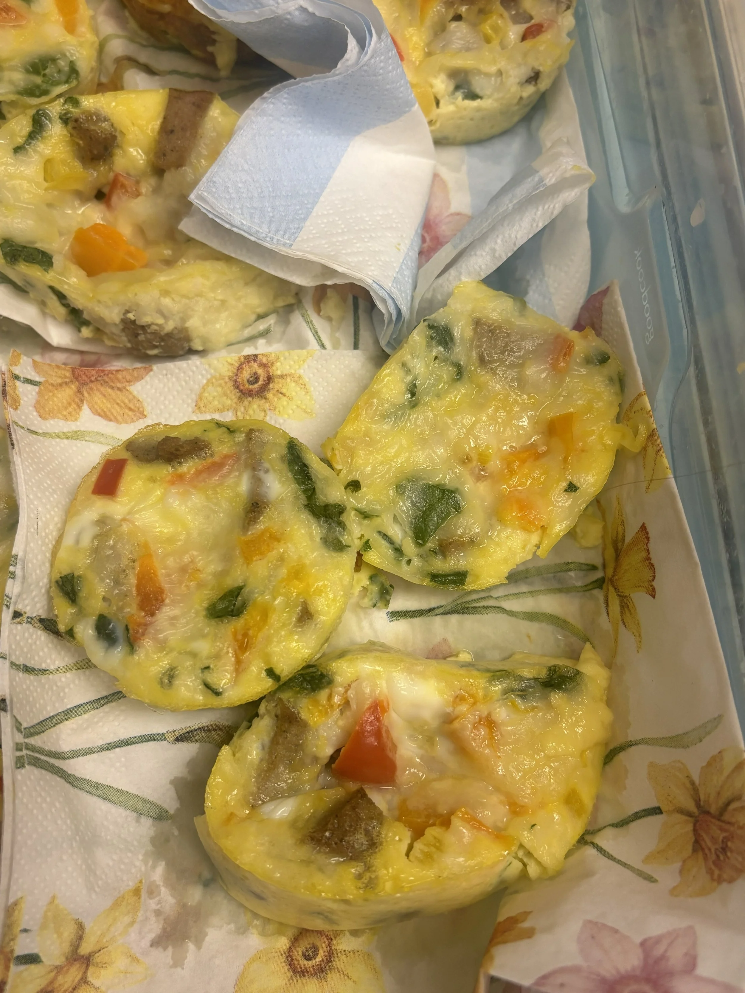  A close-up of mini egg muffins with colorful vegetables and cheese on floral napkins inside a container are shown. 