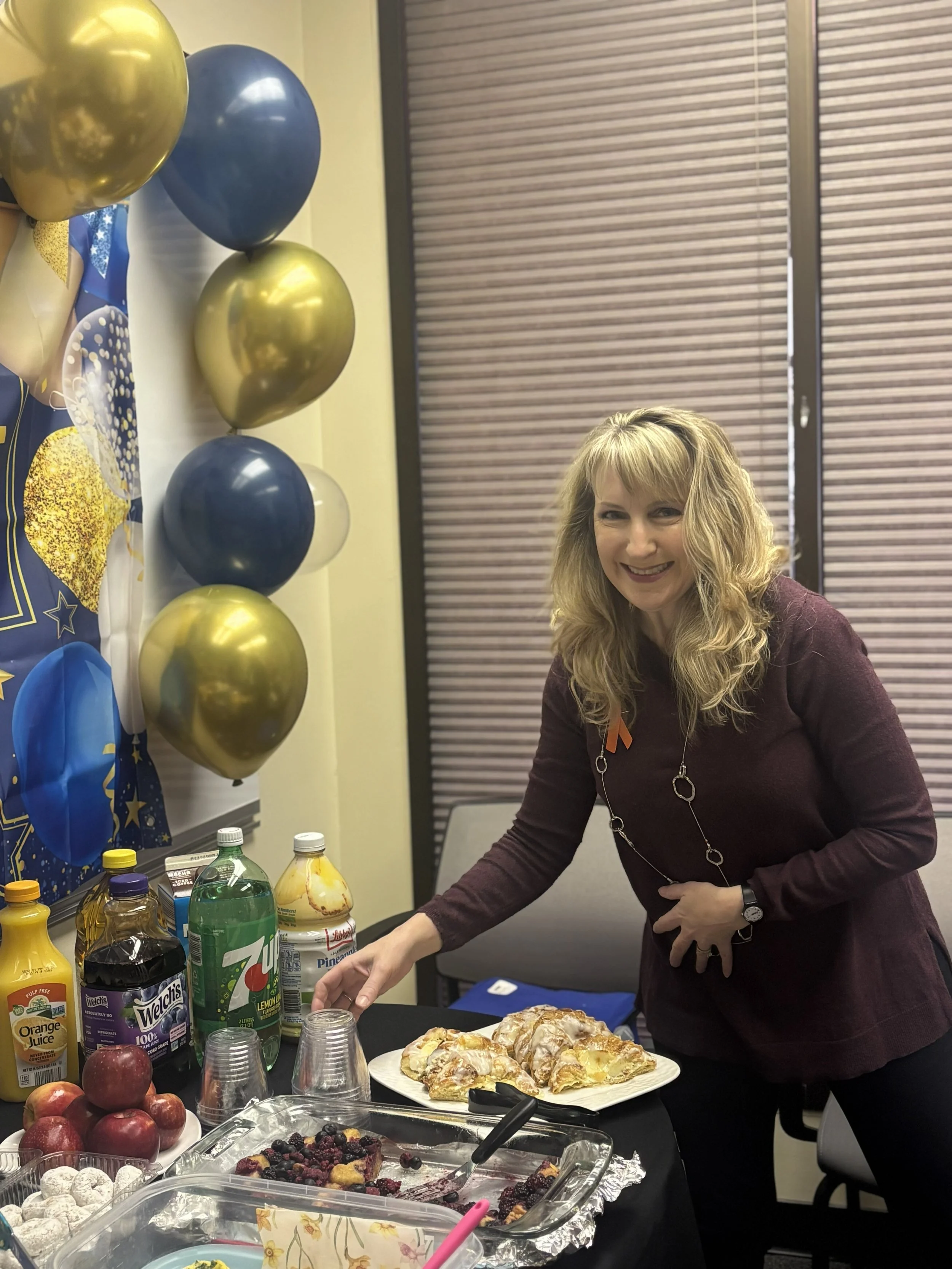  A middle aged woman is shown placing cups down onto a table. The woman is holding her necklace in place with her other hand so it doesn't get hang over the food. 