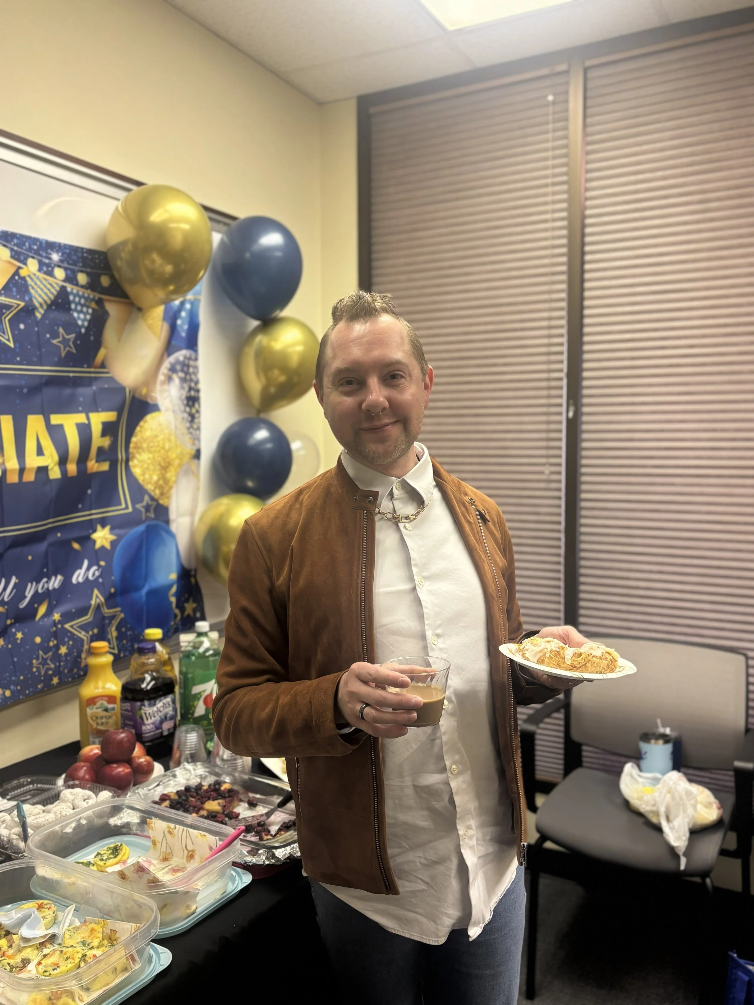  A man in a brown jacket and white button up shirt is shown holding a plate of food in one hand and a cup of coffee in his other hand. Behind him is the spread of breakfast foods and drinks. 
