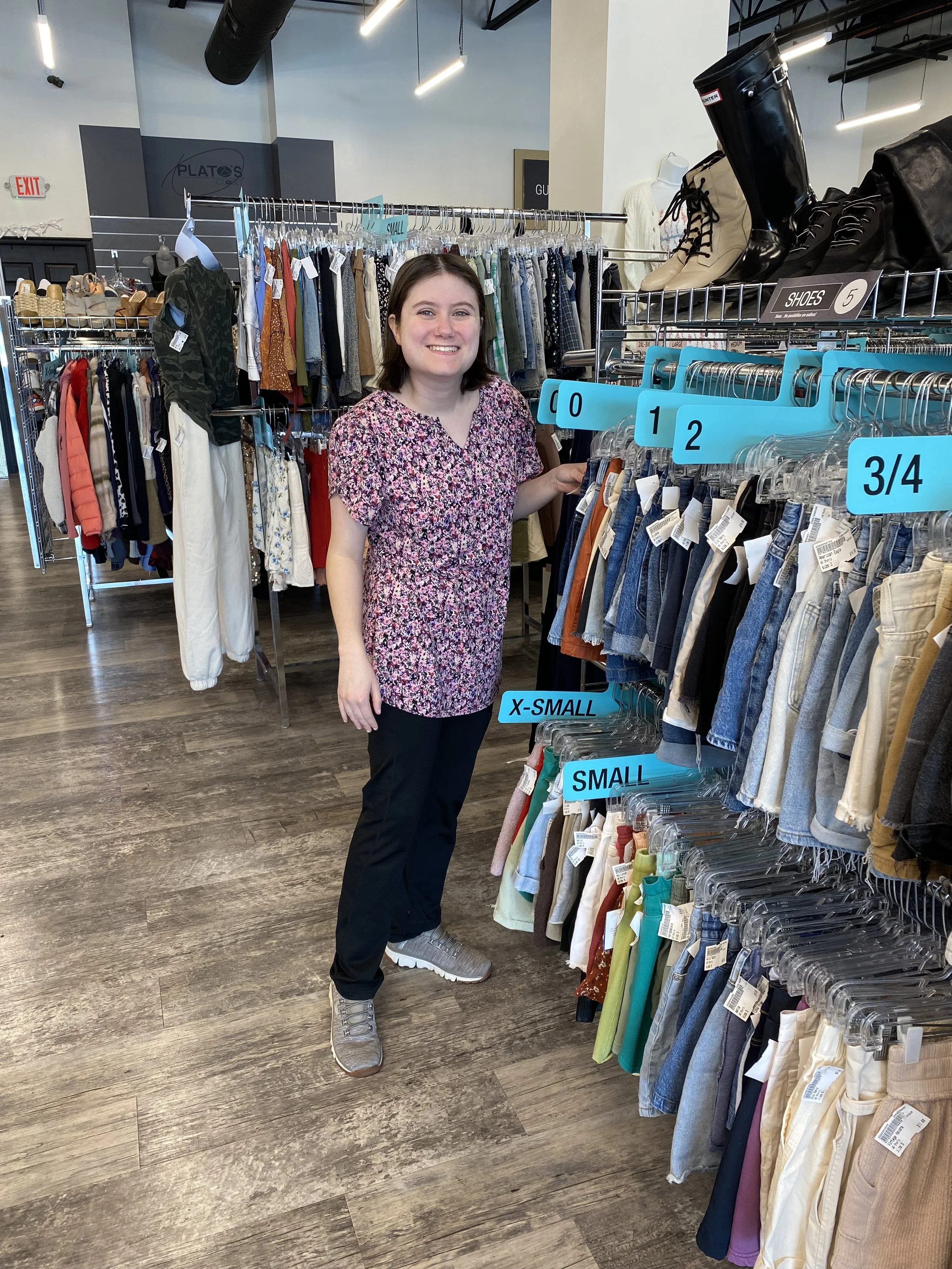  A woman is a pink floral blouse and black pants is shown standing next to a rack of women's pants in varying sizes at Plato's Closet. She is pausing from organizing the rack.  
