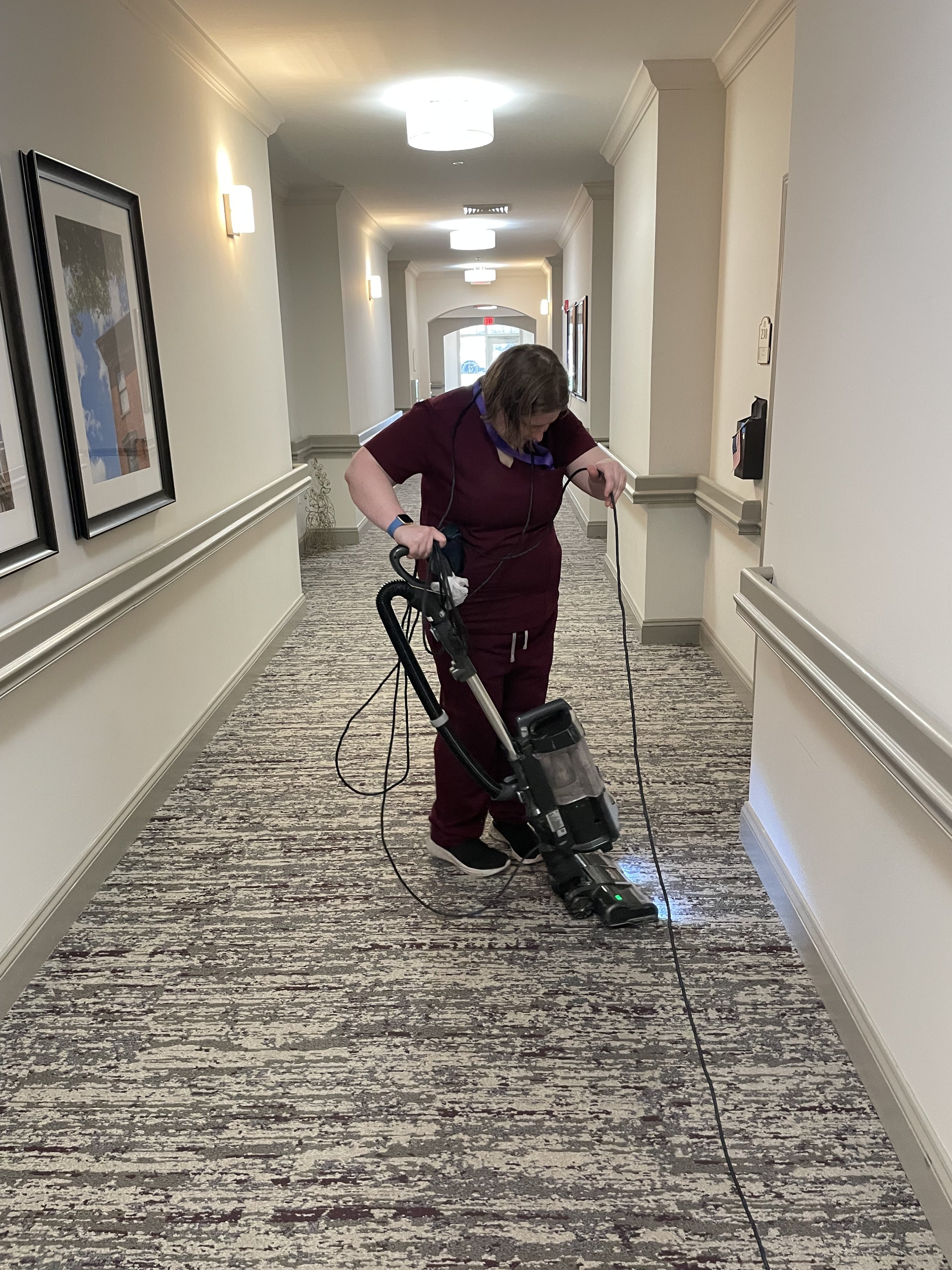  A woman is shown vacuuming a patterned carpet in a hallway. The woman is looking down at the carpet as she vacuums and is holding the cord in her free hand. The woman is wearing black pants and a maroon scrub shirt. She has dark blonde hair that is 