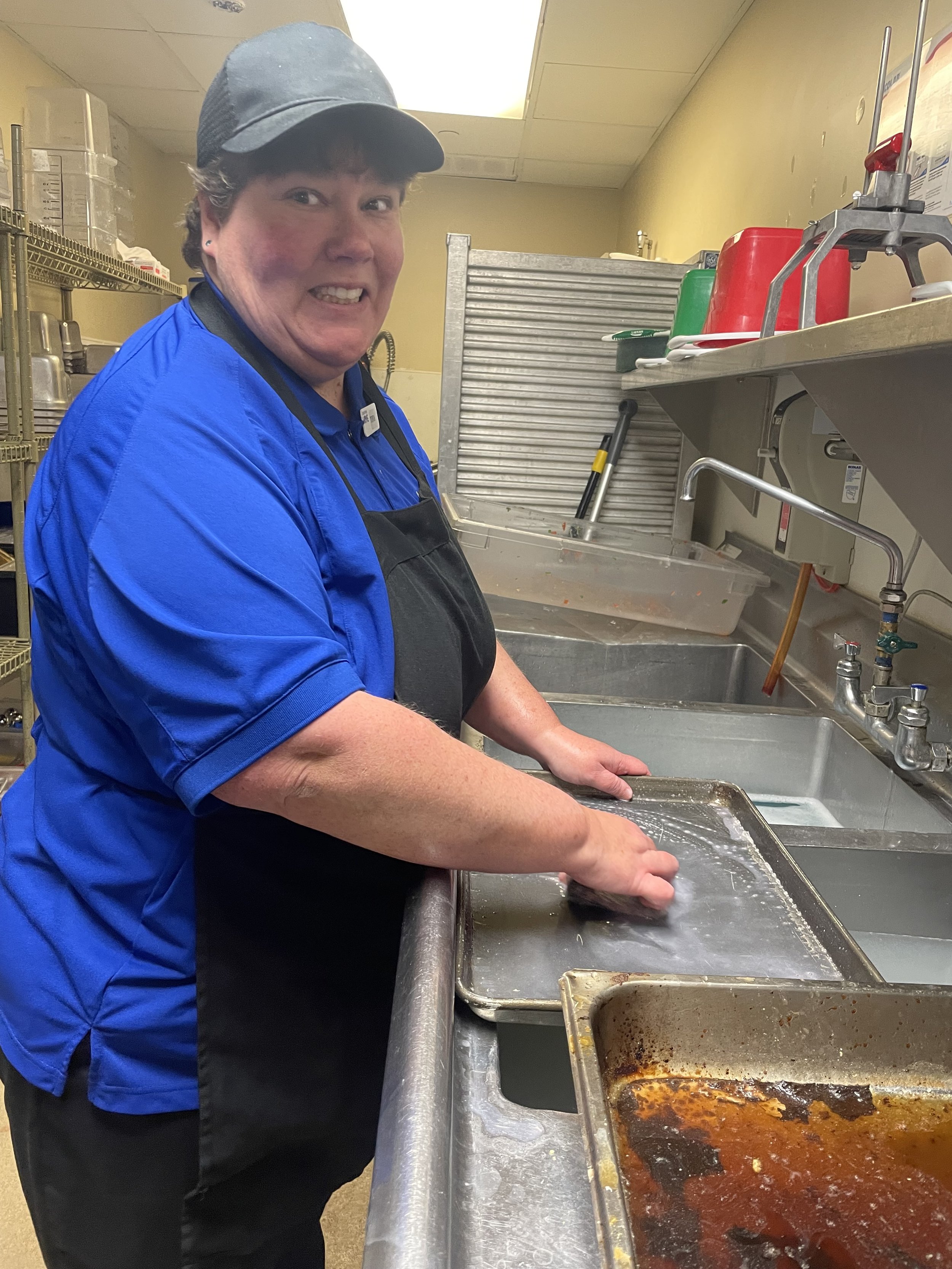  A woman is shown scrubbing a pan with soap. Both the woman's hands are on the pan, and she is looking at the camera and offering a smile. The woman is wearing dark pants, a royal blue polo shirt, a black apron and a black hat. The kitchen and sink a