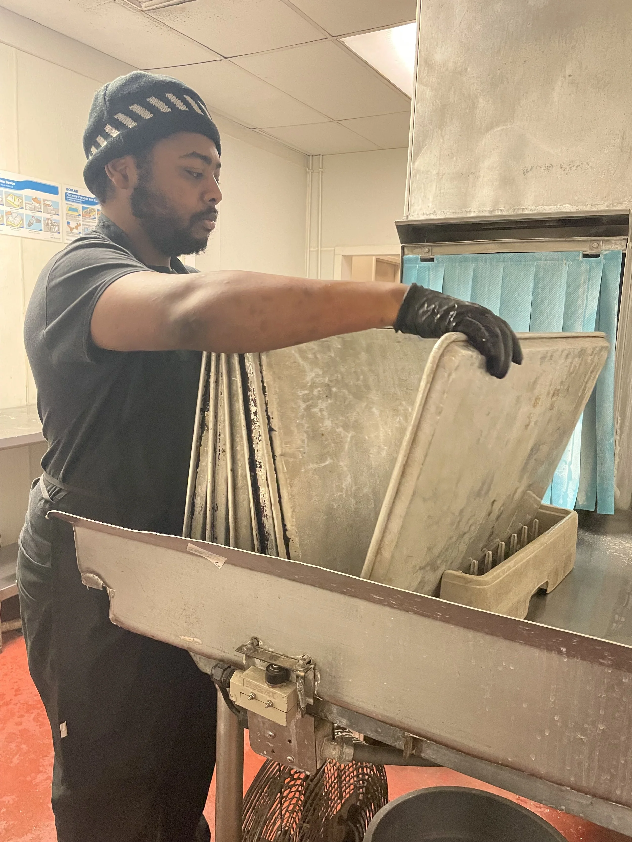  A Black man with dark clothes and a black beanie on is shown loading an industrial dishwasher bin with metal trays. The dish room is slightly shown in the background behind the man, and the dishwasher is closest to the man as he is loading the bin w