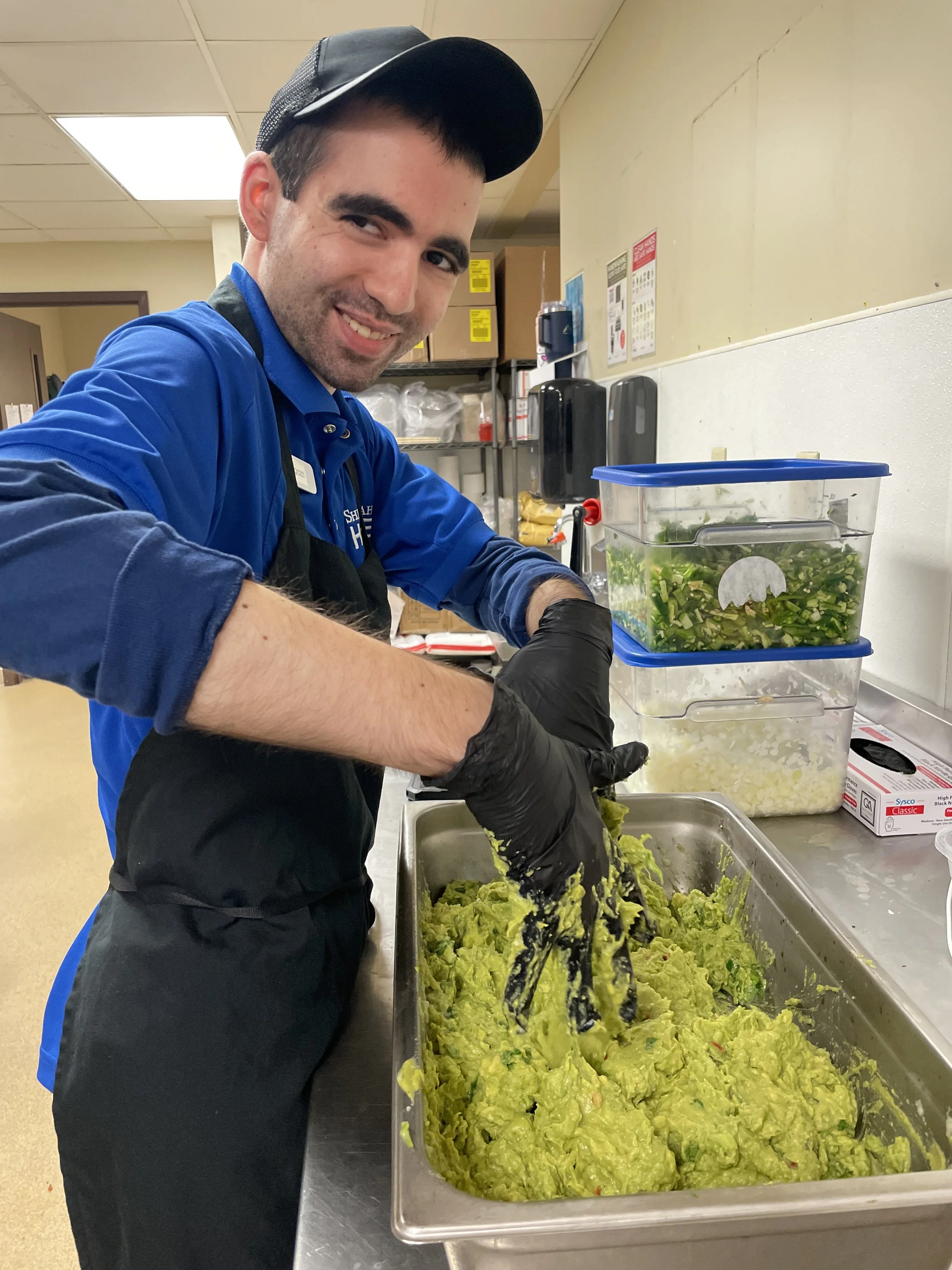  A young man is shown smiling at the camera as he makes guacamole. The man has his hands slightly in the tray of guacamole and is mixing it up. The man is wearing a royal blue polo shirt with another blue 3/4 length shirt layered below it. He is also