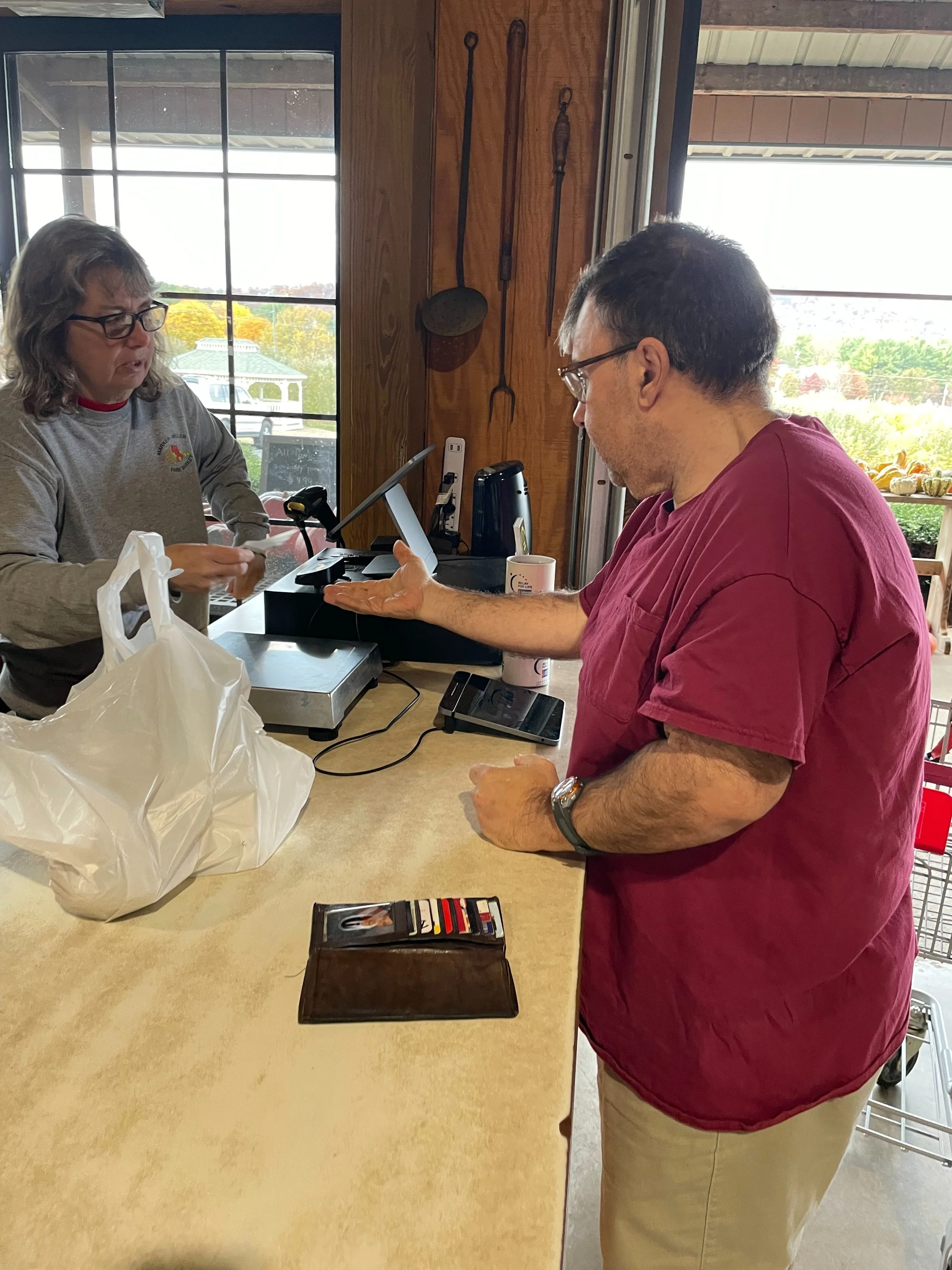 A middle aged man is shown with his hand out waiting for his change at a cash register. The man's wallet is beside him, and the grocery bag holding his purchases is near the cashier who is checking him out.