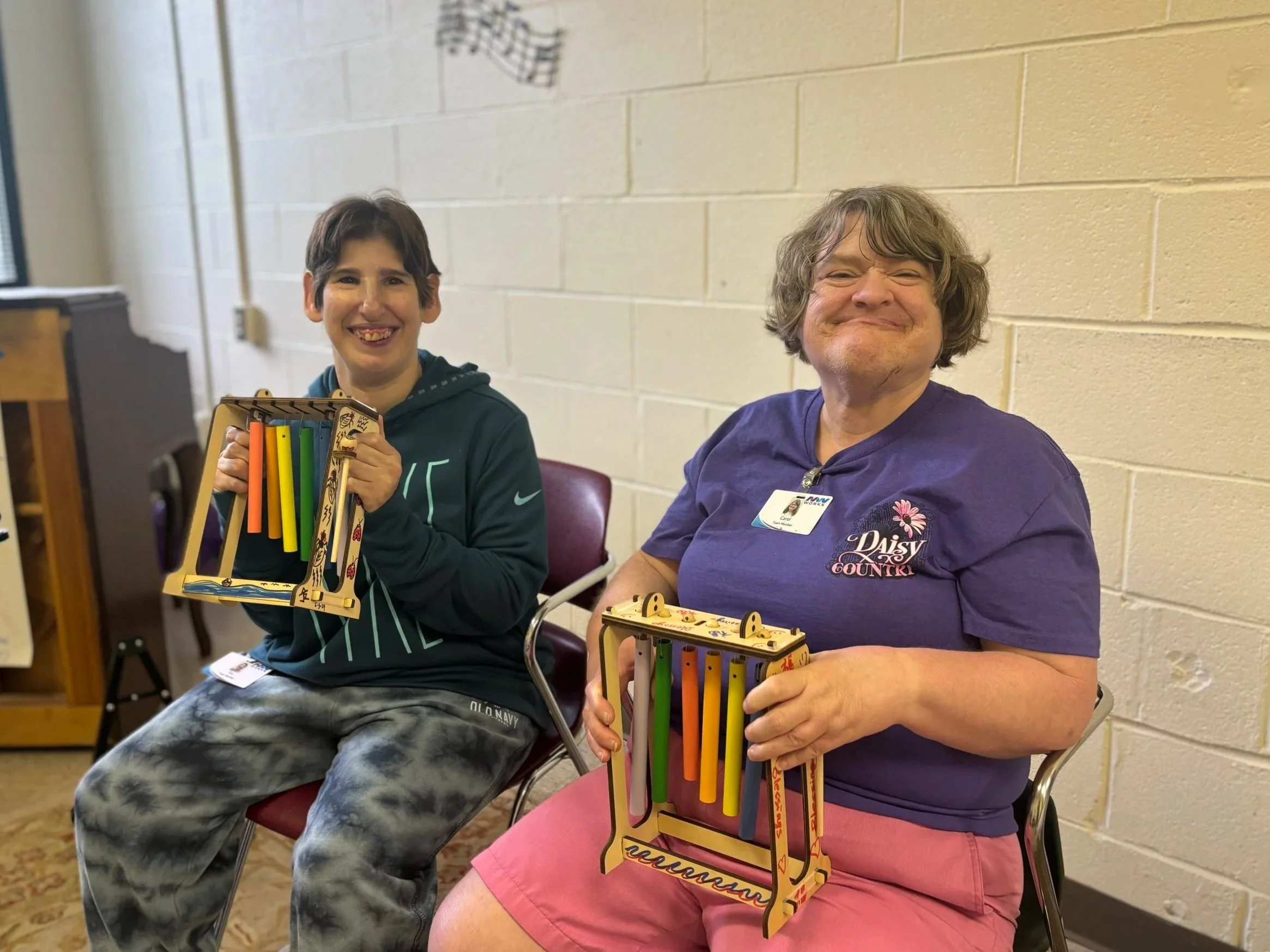Two women smile at the camera as they hold up colorful chimes. Both women are participating in music therapy at NW Works.