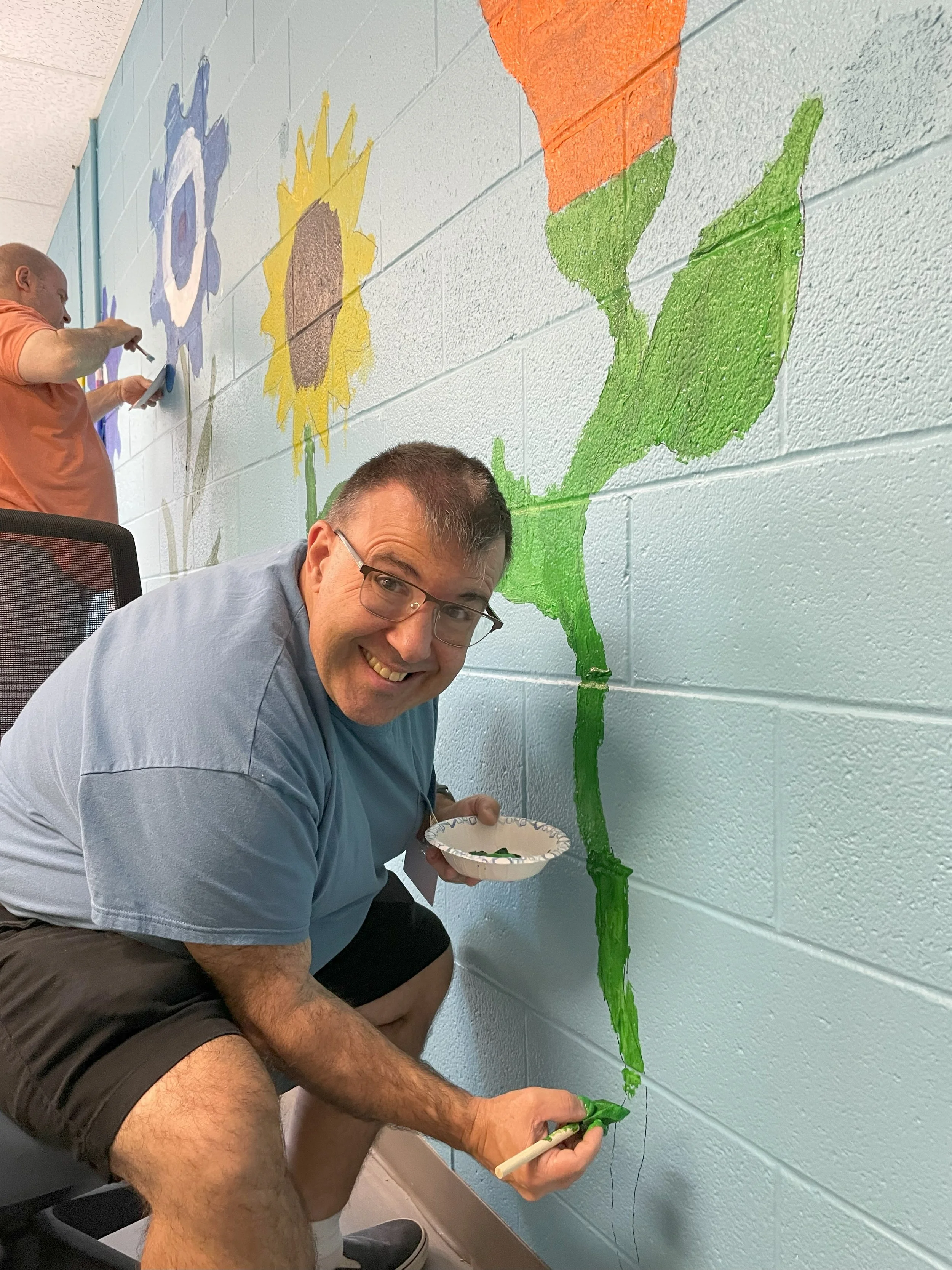 A man is shown smiling at the camera as he pauses from painting. The man is painting the wall and flowers inside the Digital Literacy Lab at NW Works.