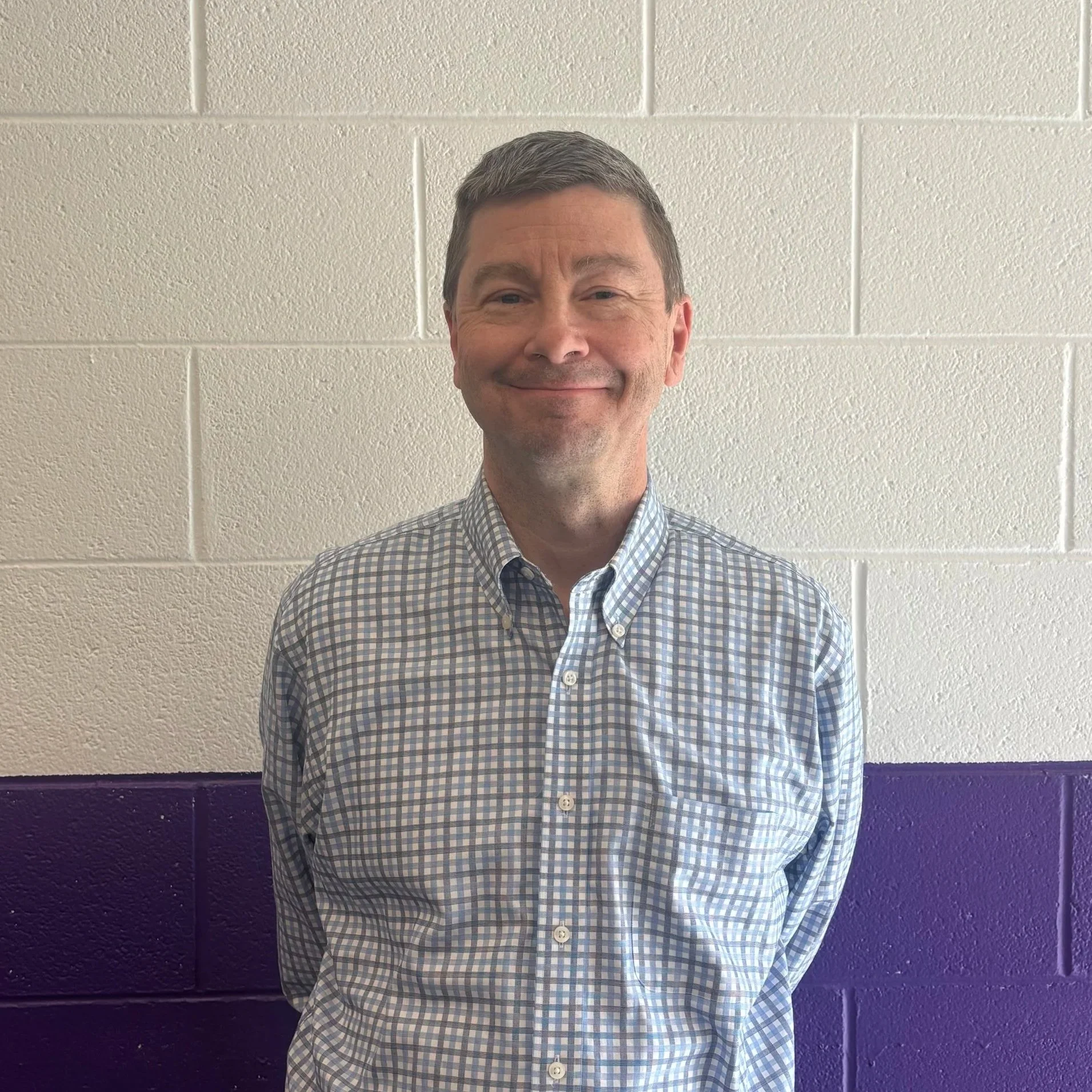 A middle aged man, Todd, is shown smiling for a photo as he stands against a white and purple brick wall. Todd is a Caucasian man with brown hair and blue eyes.  He is wearing a blue, white, and grey checkered button up shirt.