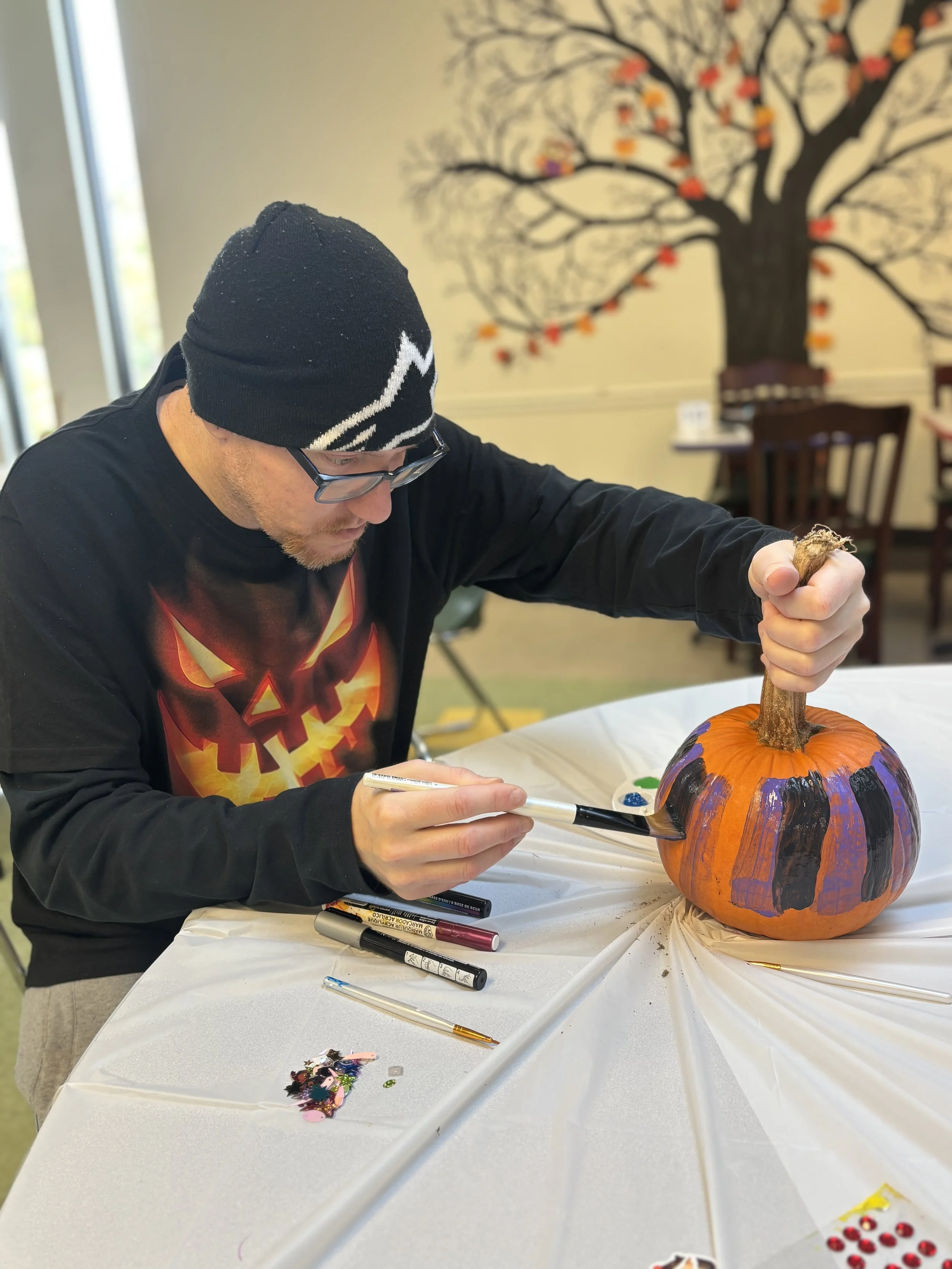 A young man is shown sitting at a table as he paints a pumpkin. He is painting the pumpkin with black and purple alternating stripes. The young man is wearing a long sleeve jack'o-lantern graphic shirt, a beanie, and glasses.