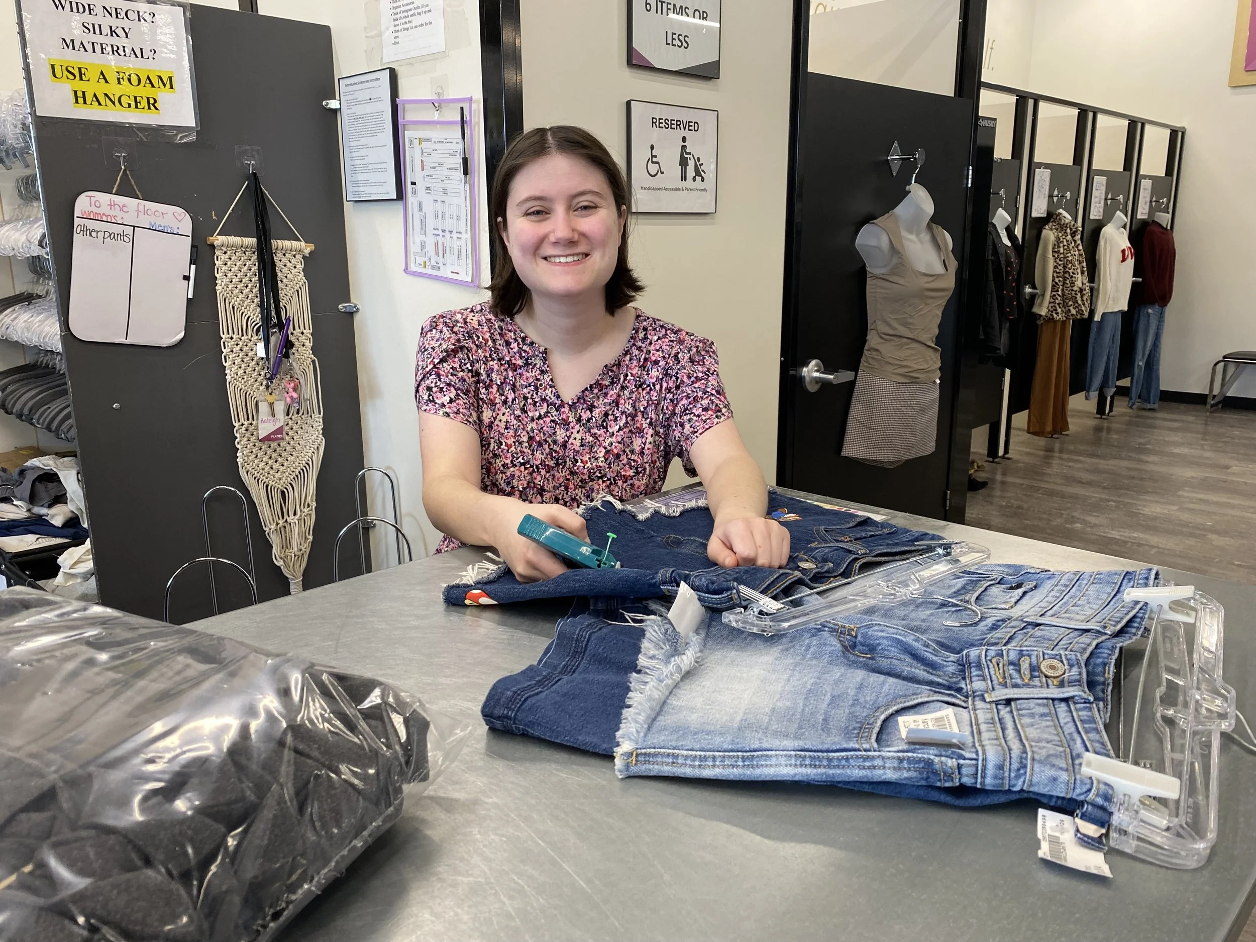 A young woman is shown smiling at the camera as she pauses from tagging shorts. She has several pairs of shorts in front of her that have already been tagged. Beside her are fitting rooms.